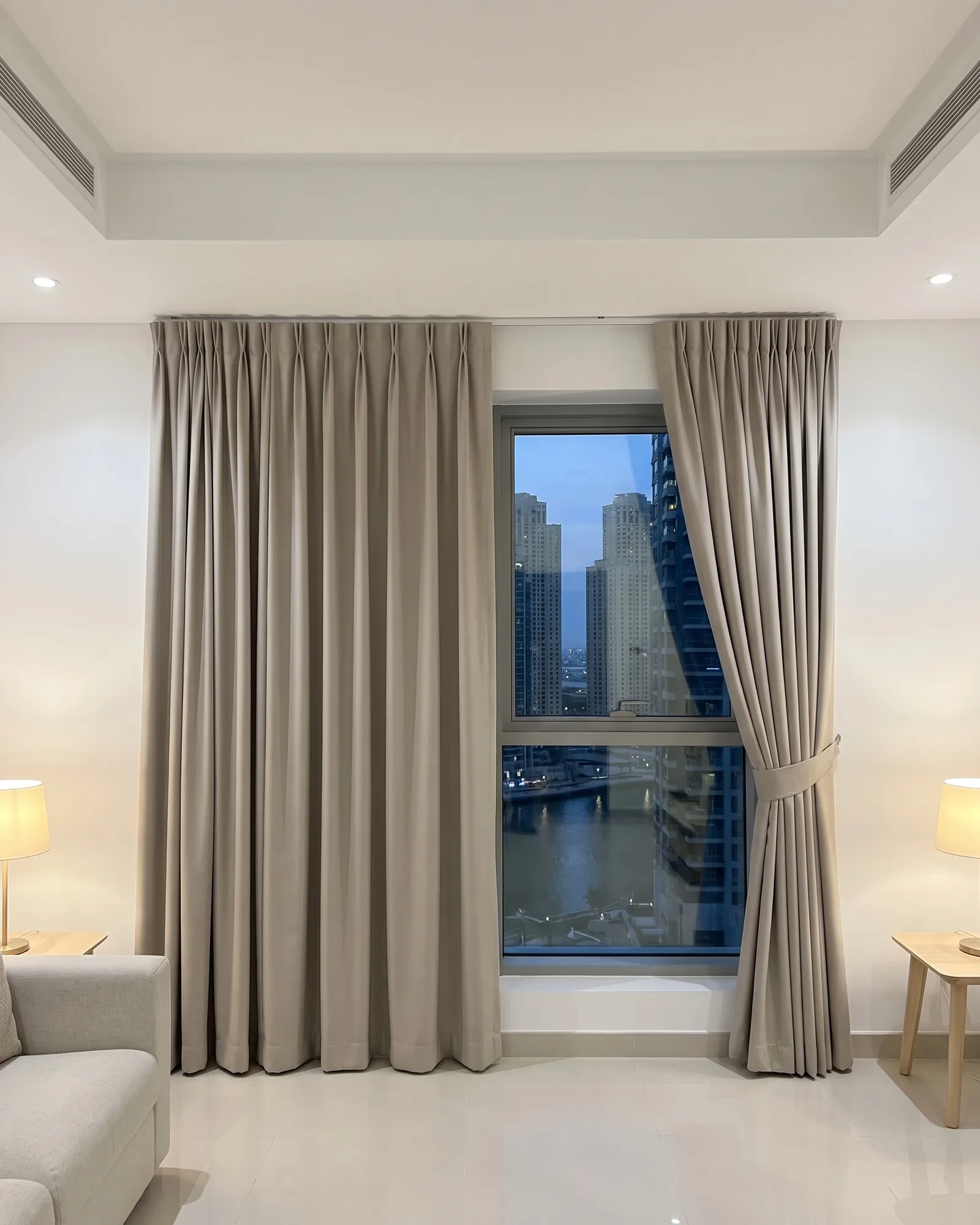 Greige pinch pleat curtains in a living room at dusk, one half closed and one tied back, the marina through the window, lamps lit