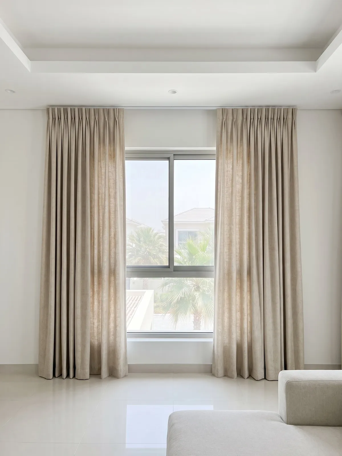 Oatmeal linen pinch pleat curtains drawn to both sides of a living-room window, palms through the glass, a pale sofa in front
