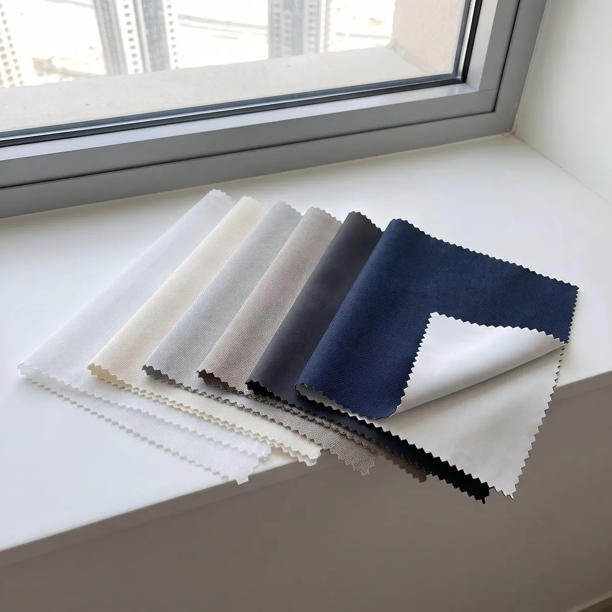 Curtain fabric samples fanned on a sunlit sill, white voile, ivory, light grey, greige, charcoal and navy turned up at a corner
