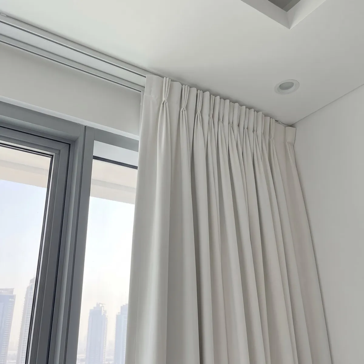 White pinch pleat curtain drawn into a bunch at the end of a ceiling track beside a window, its heading hiding the track