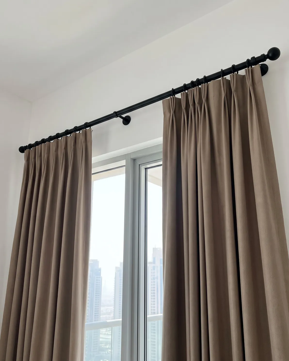Taupe pinch pleat curtains hooked into the rings of a black pole, drawn to both sides of a glass door, seen from below at an angle