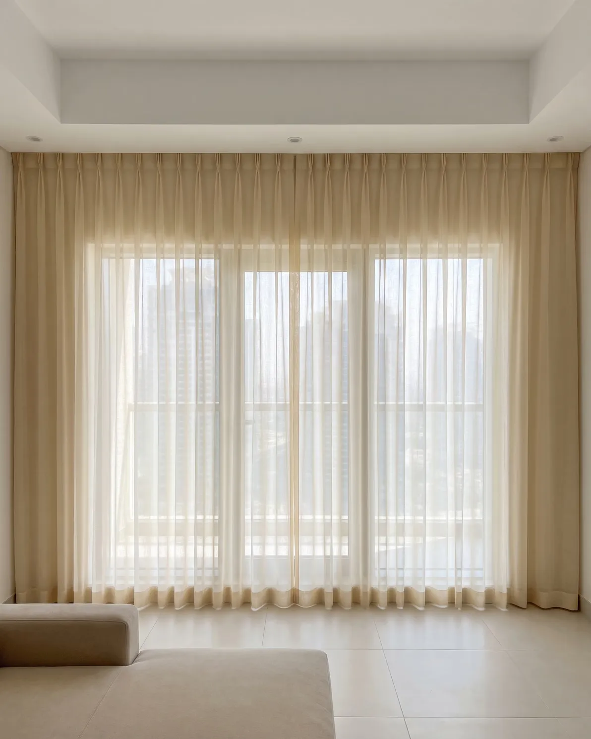 Cream voile pinch pleat sheer closed across wide living-room glass, daylight glowing through it, towers soft behind, a sofa in front