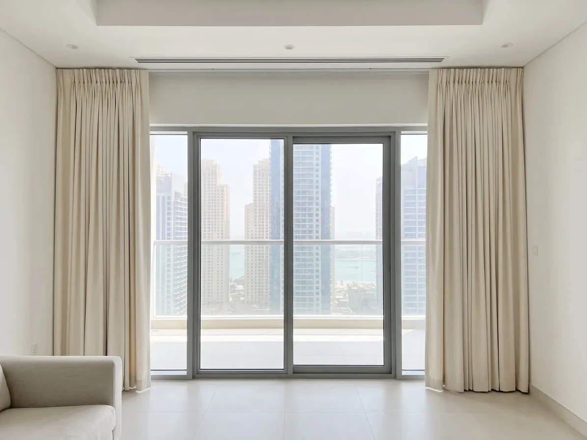 Ivory pinch pleat curtains drawn open on a long ceiling track beside a wide sliding door, towers and the sea beyond, an armchair