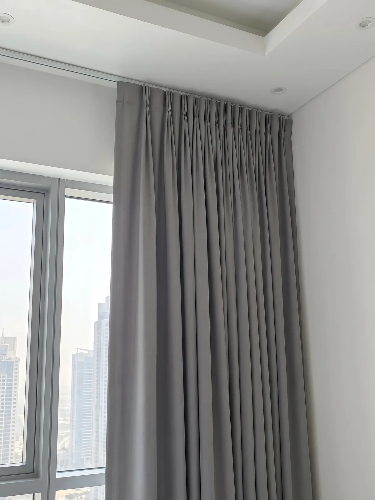Mid grey pinch pleat curtain closed under a ceiling track, seen at an angle so the flat gaps between its pleats show, towers beside it
