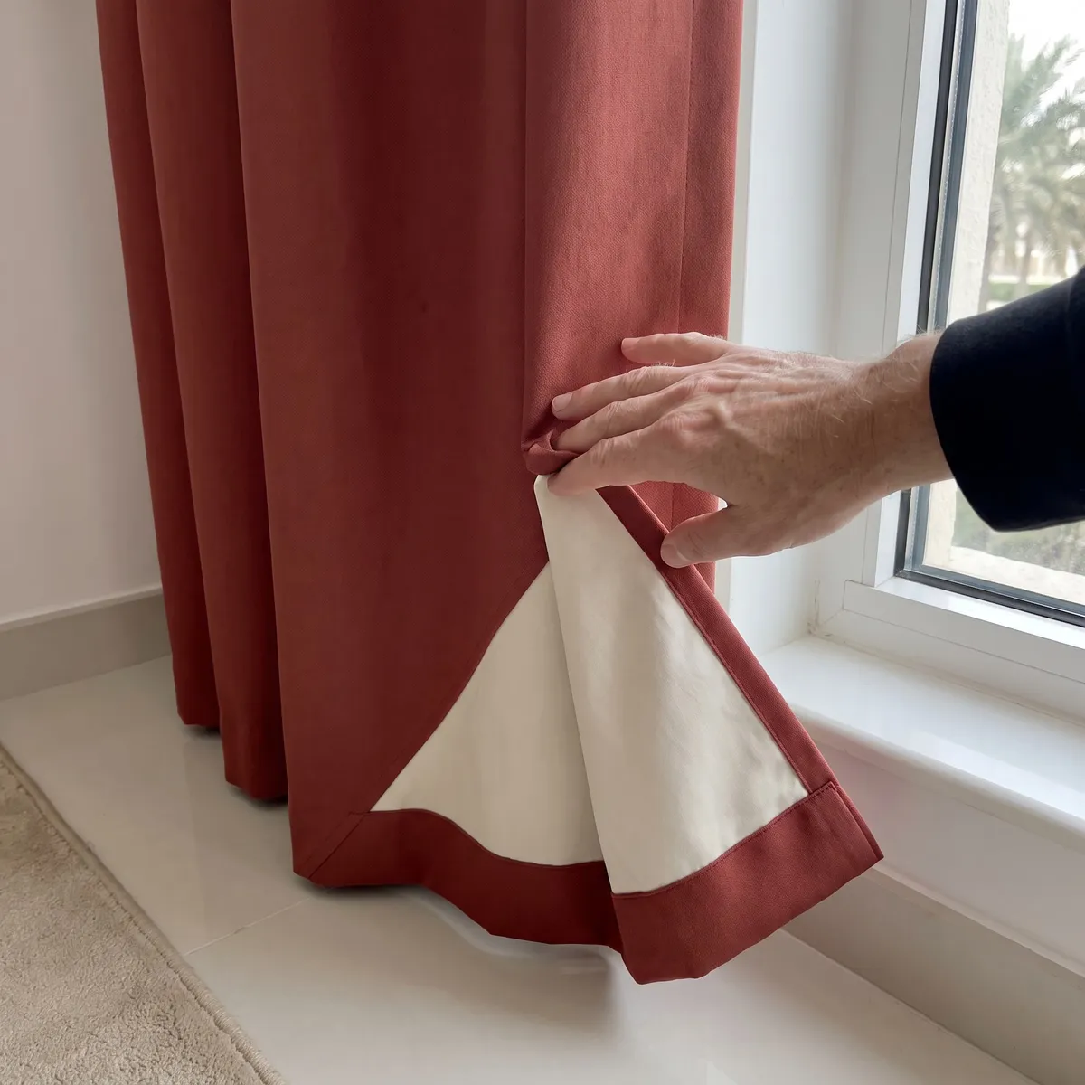 A hand folding back the bottom corner of a brick-red curtain to show its ivory lining stopping above the red hem band