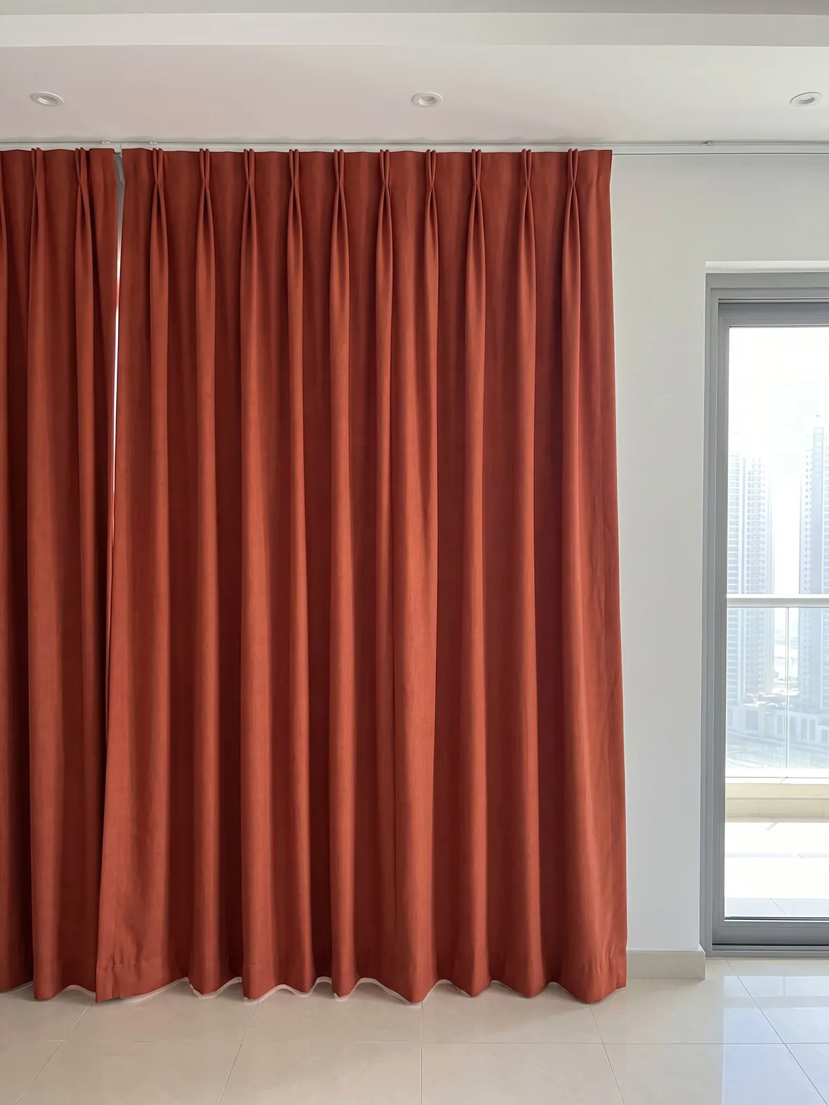 Rust-red pinch-pleat curtains on a white track under the ceiling, small neat pleats, smooth folds, a bare balcony door with towers at right