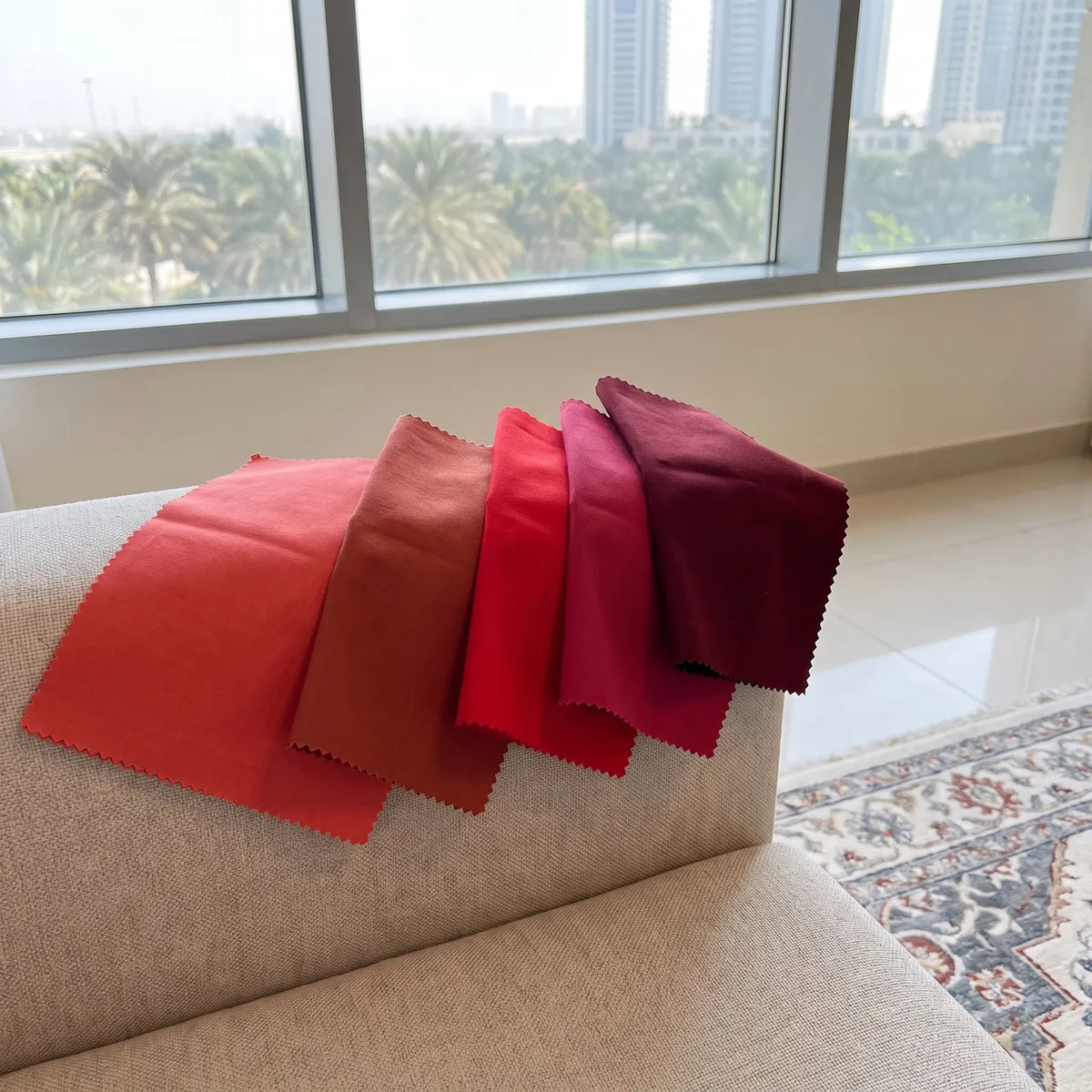 Five red fabric swatches with zigzag edges fanned over an oatmeal sofa arm, coral to oxblood, a patterned rug and a window with palms beyond