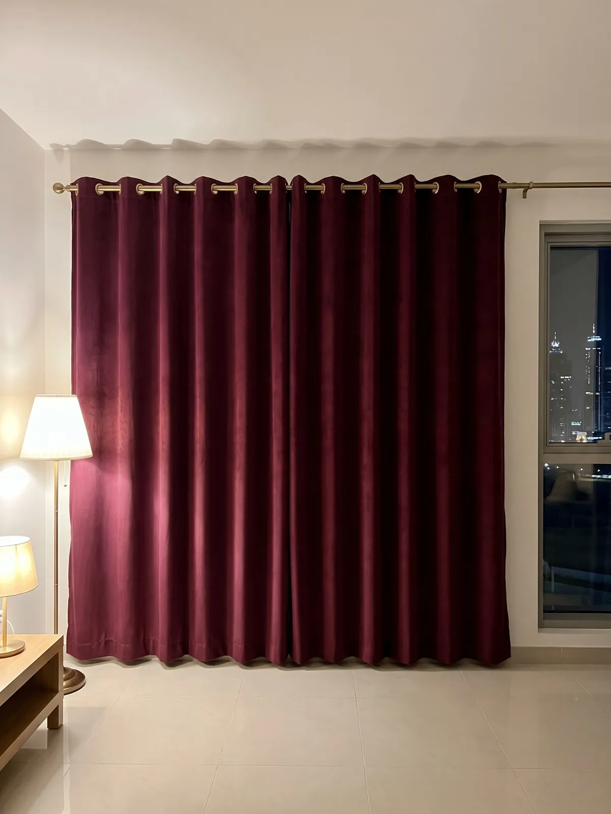 Wine-red eyelet curtains closed on a brass pole at night, a lit floor lamp and table lamp at left, a window with lit towers at right
