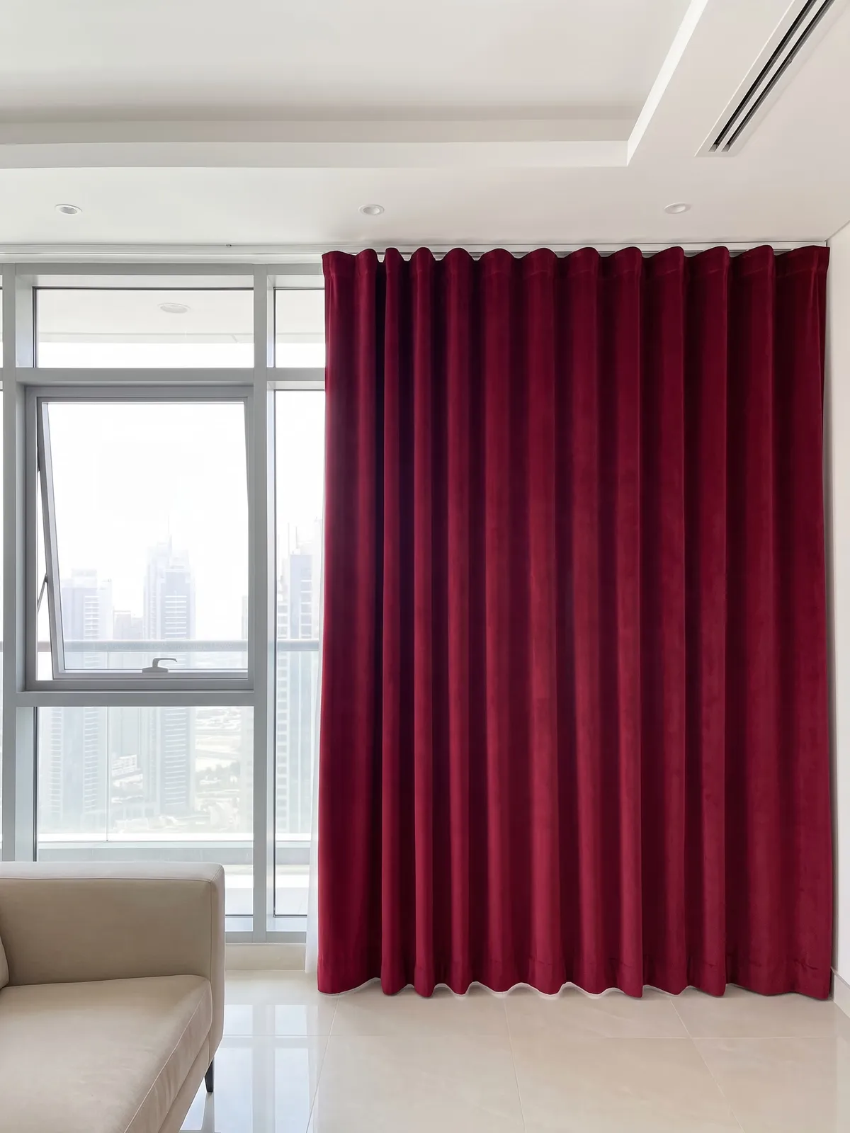 Ruby-red velvet wave curtain on a white ceiling track beside a tall window with a tower view, one even red, a sofa at lower left