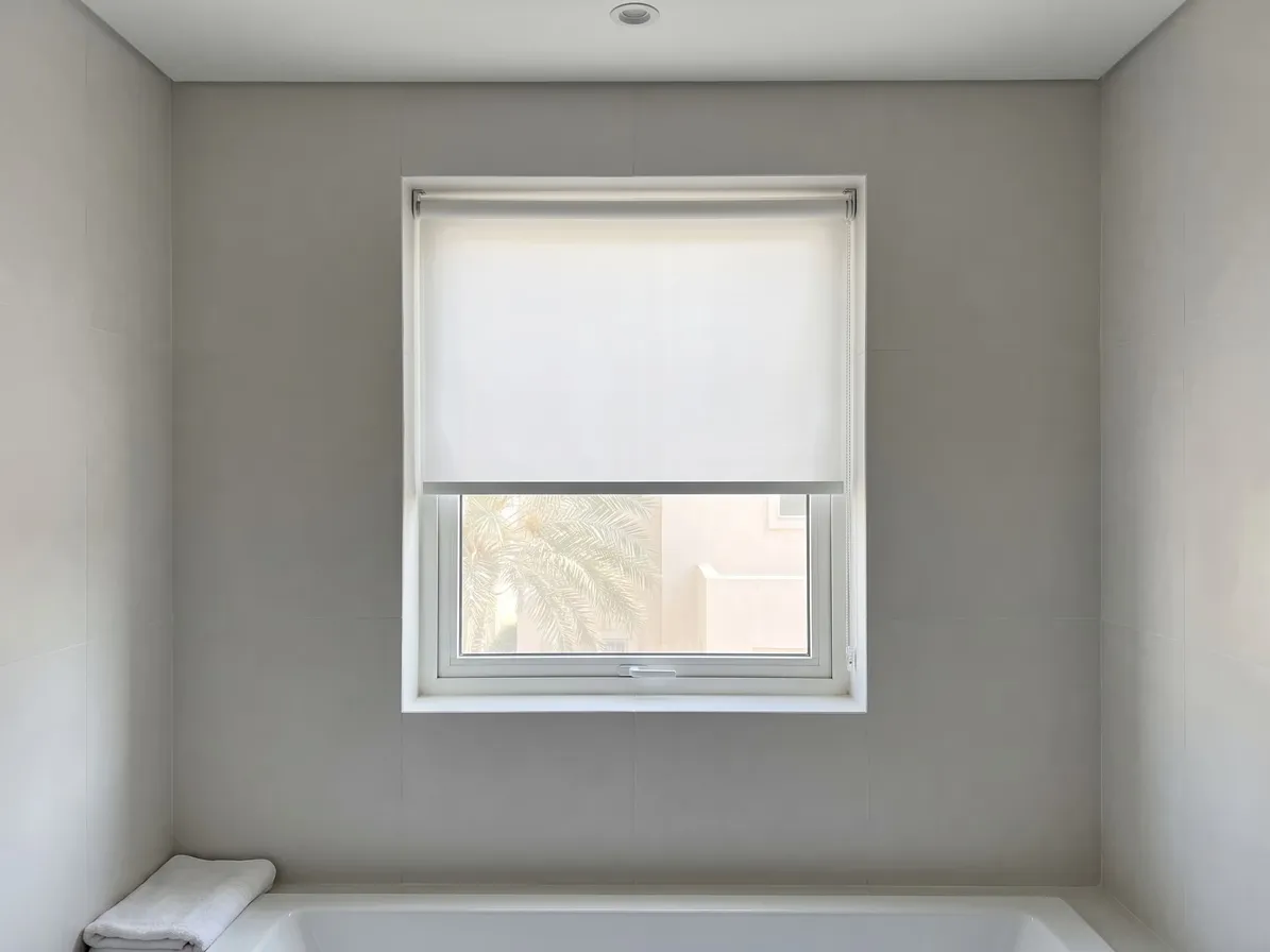 White roller blind half lowered in a small window in a tiled bathroom wall, a bead chain at right, a palm outside, a towel on the bath edge