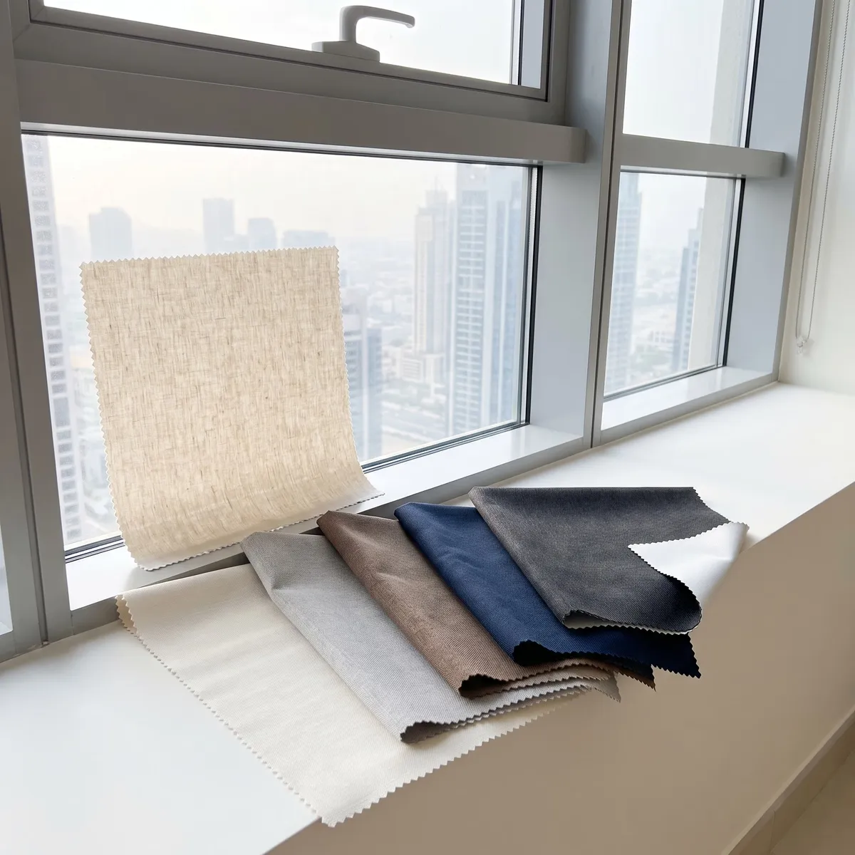 Six fabric samples with zigzag edges on a white window sill, natural linen against the glass, then ivory, grey, mocha, navy and charcoal