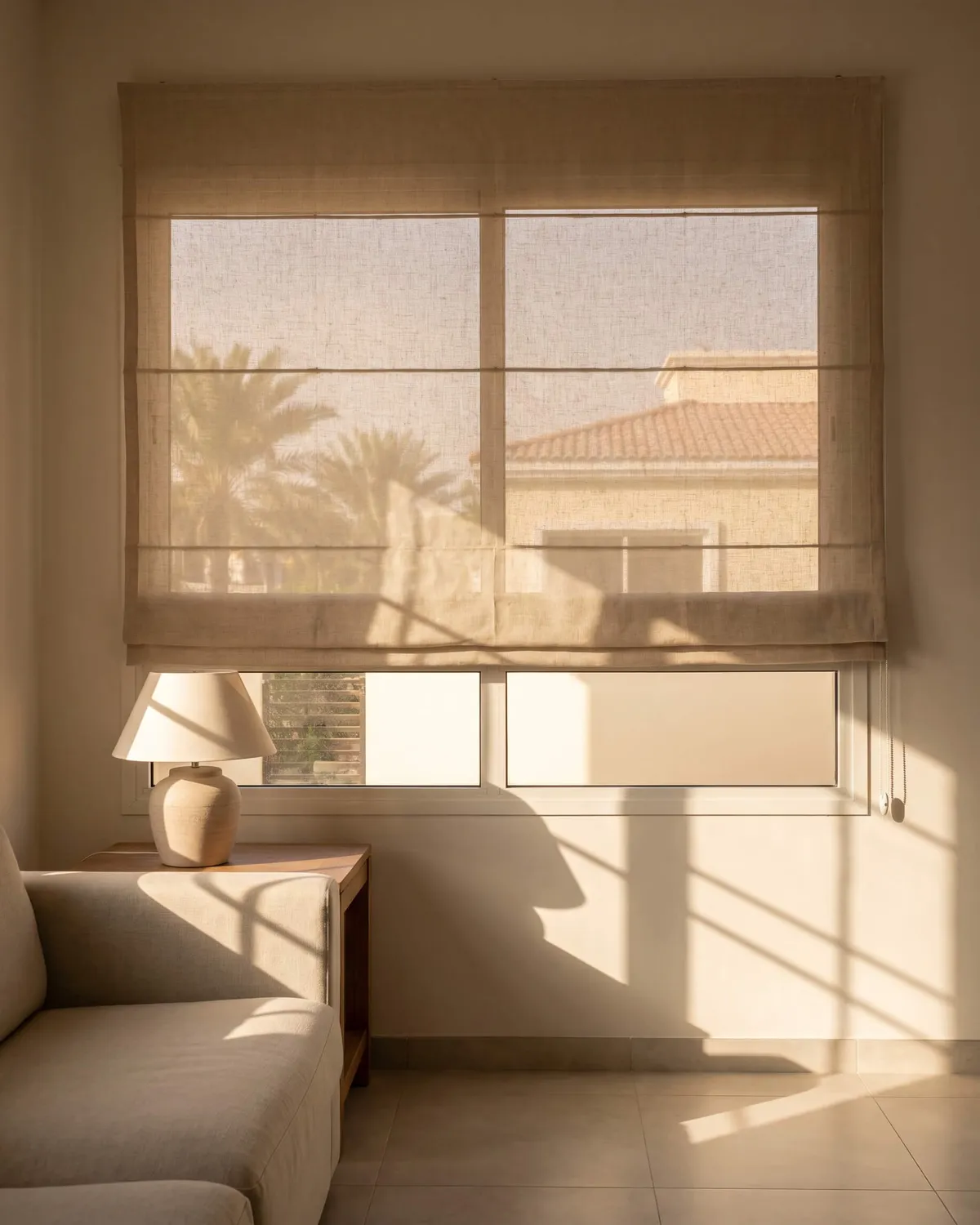 Linen Roman blind lowered most of the way over a living-room window, afternoon sun glowing through