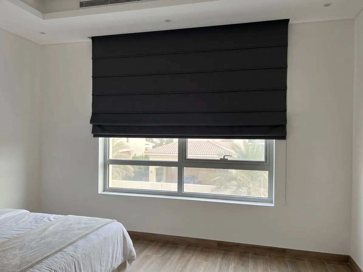 Charcoal Roman blind lowered most of the way over a bedroom window, villa roofs and palms in the glass below, a made bed at lower left