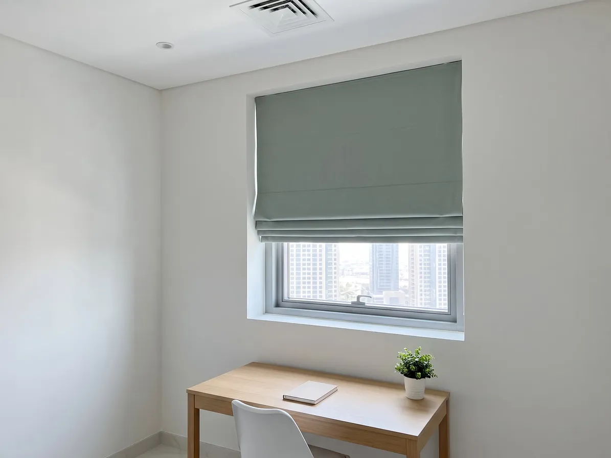 Sage-green Roman blind half raised inside a small window recess, buildings beyond the lower glass, a wooden desk with a plant below