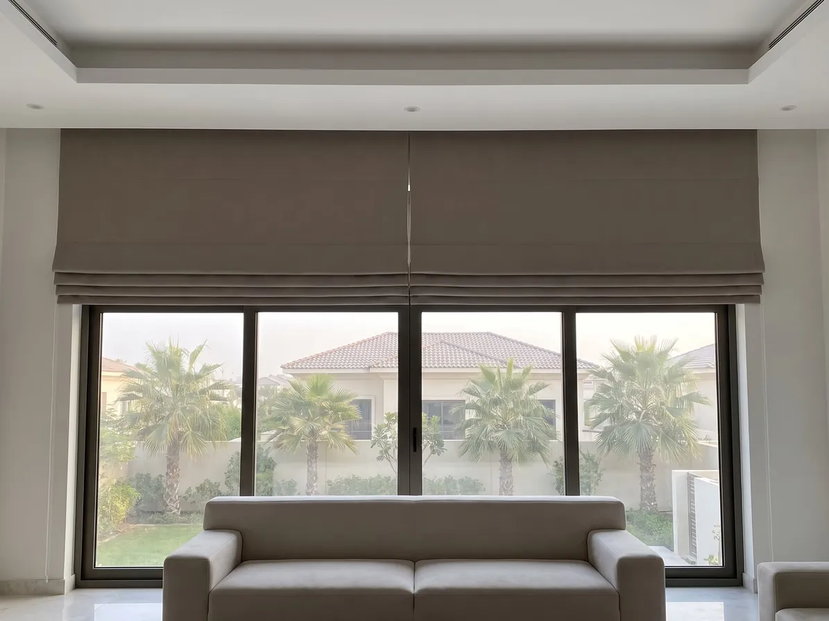 Two taupe Roman blinds side by side, raised above a wide four-pane window with palms and a villa roof outside, a cream sofa in front