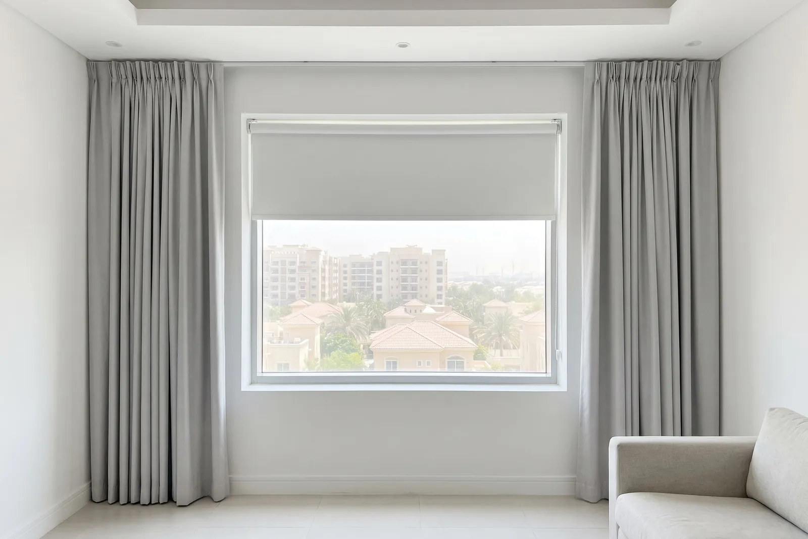 Light grey curtains open at both sides of a window with a white roller blind raised to its top third, villas and towers outside, a sofa
