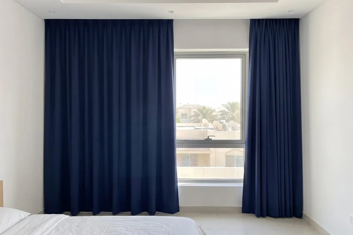 Navy lined curtains in a sunny bedroom, the left one drawn across the window and the right one open, palms and villas outside