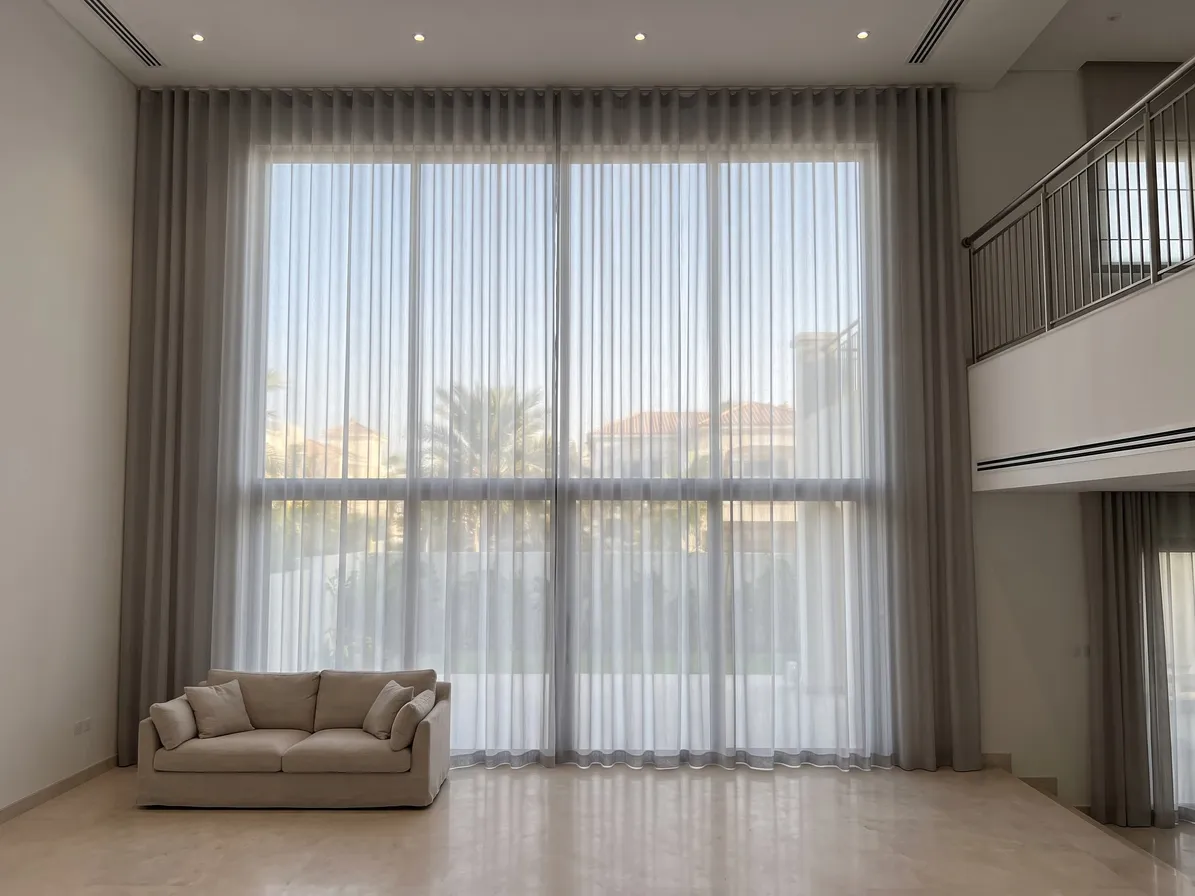 Soft grey wave-heading sheers hanging ceiling to floor over double-height glass, palms and villa roofs soft behind, a sofa below