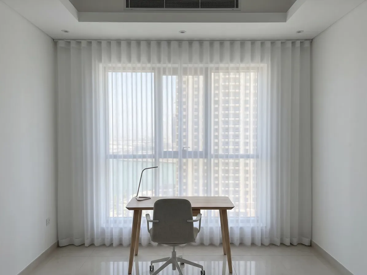White wave-heading sheer hanging past both sides of an office window, a pale wood desk and grey chair in front, towers beyond
