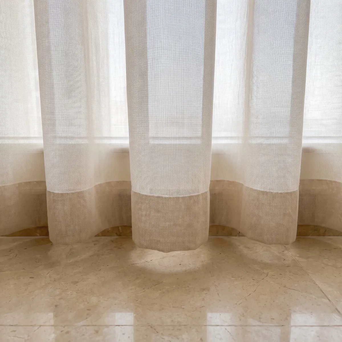 Hem of a cream sheer with a deeper sand-coloured band along the bottom, its scalloped edge hanging just above polished beige marble