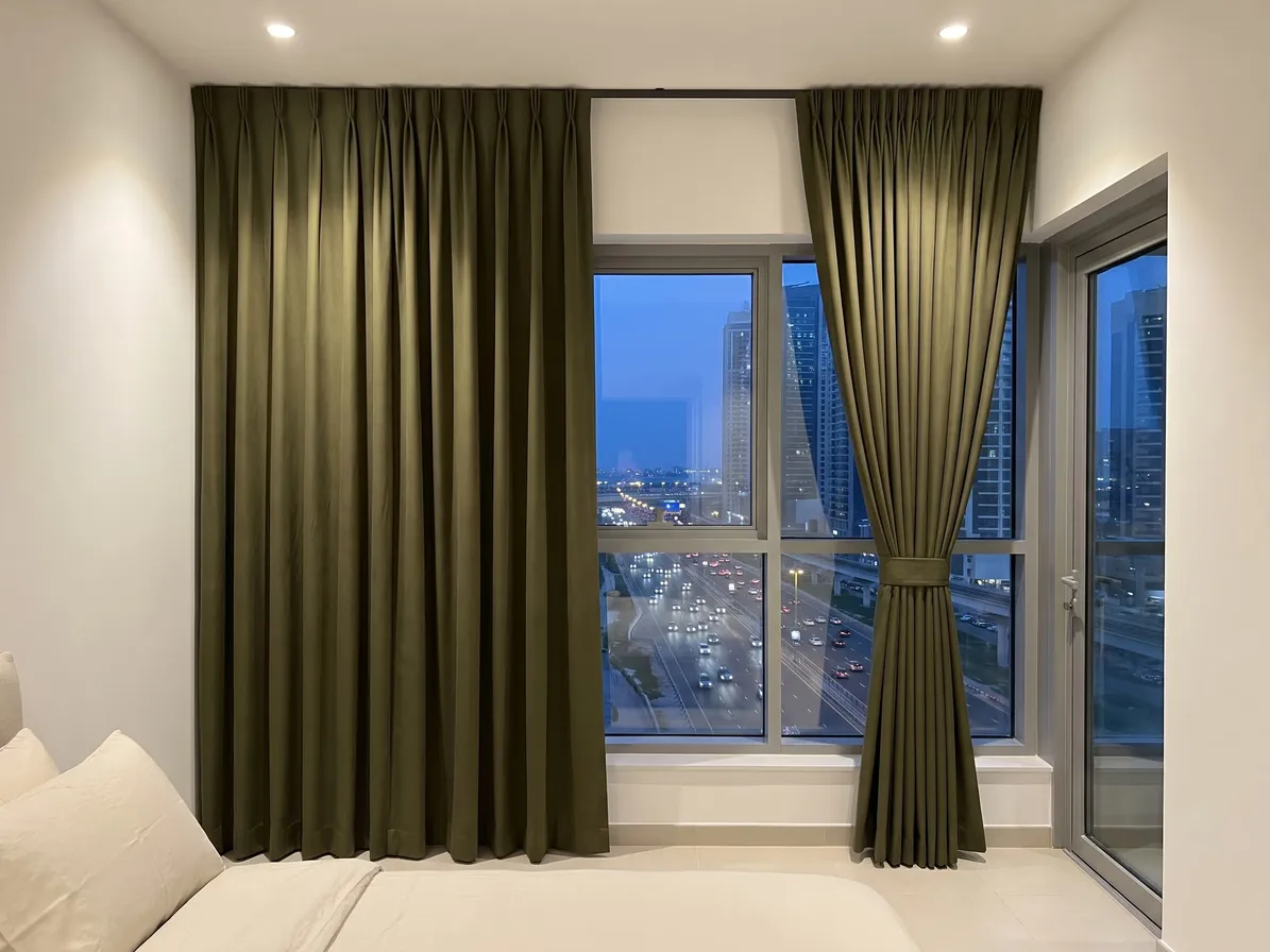 Olive heavy curtains on a bedroom window above a busy road at dusk, one half closed and one tied back, car lights on the road below