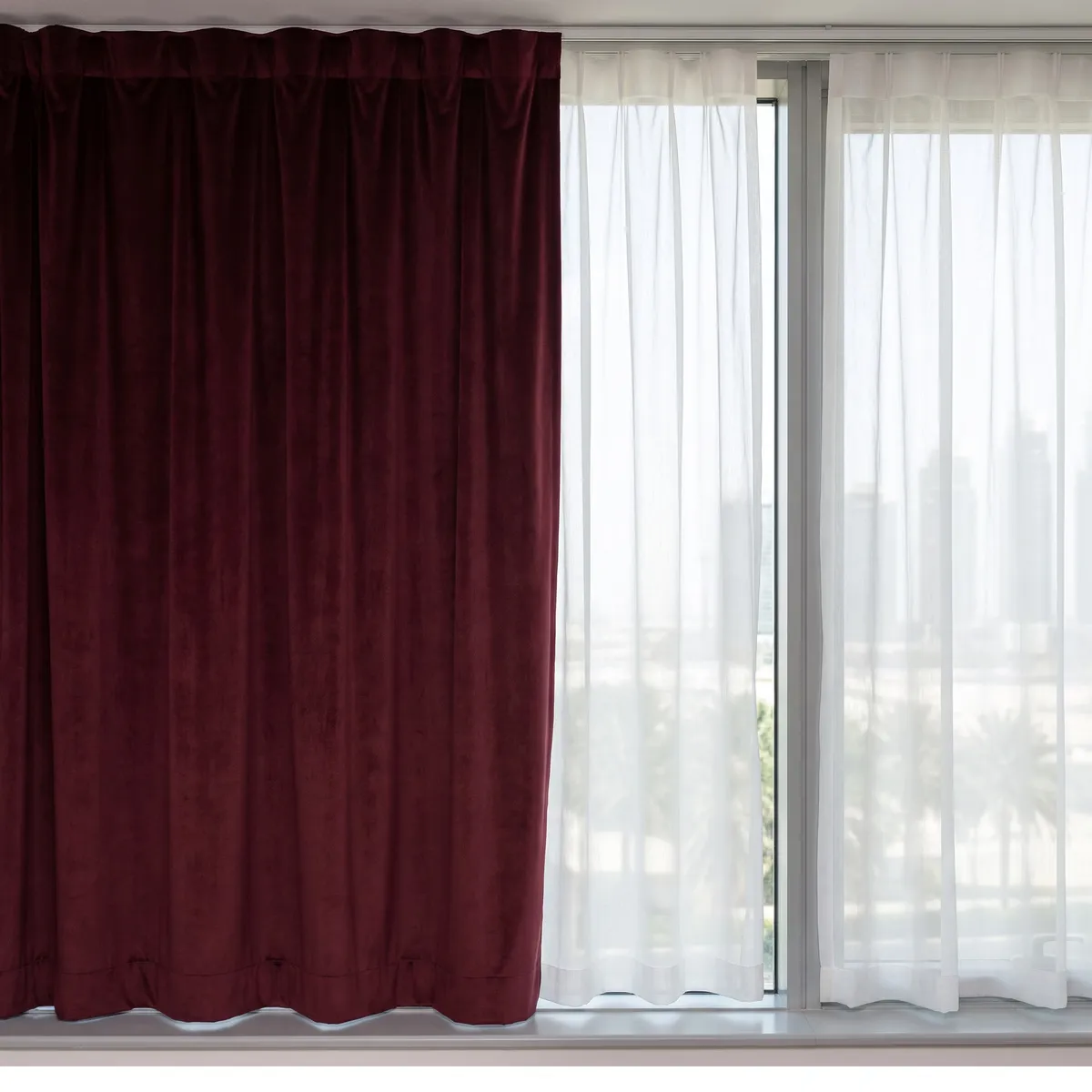 Burgundy velvet curtain beside a white voile on one window, the velvet solid and the voile glowing with towers soft through it