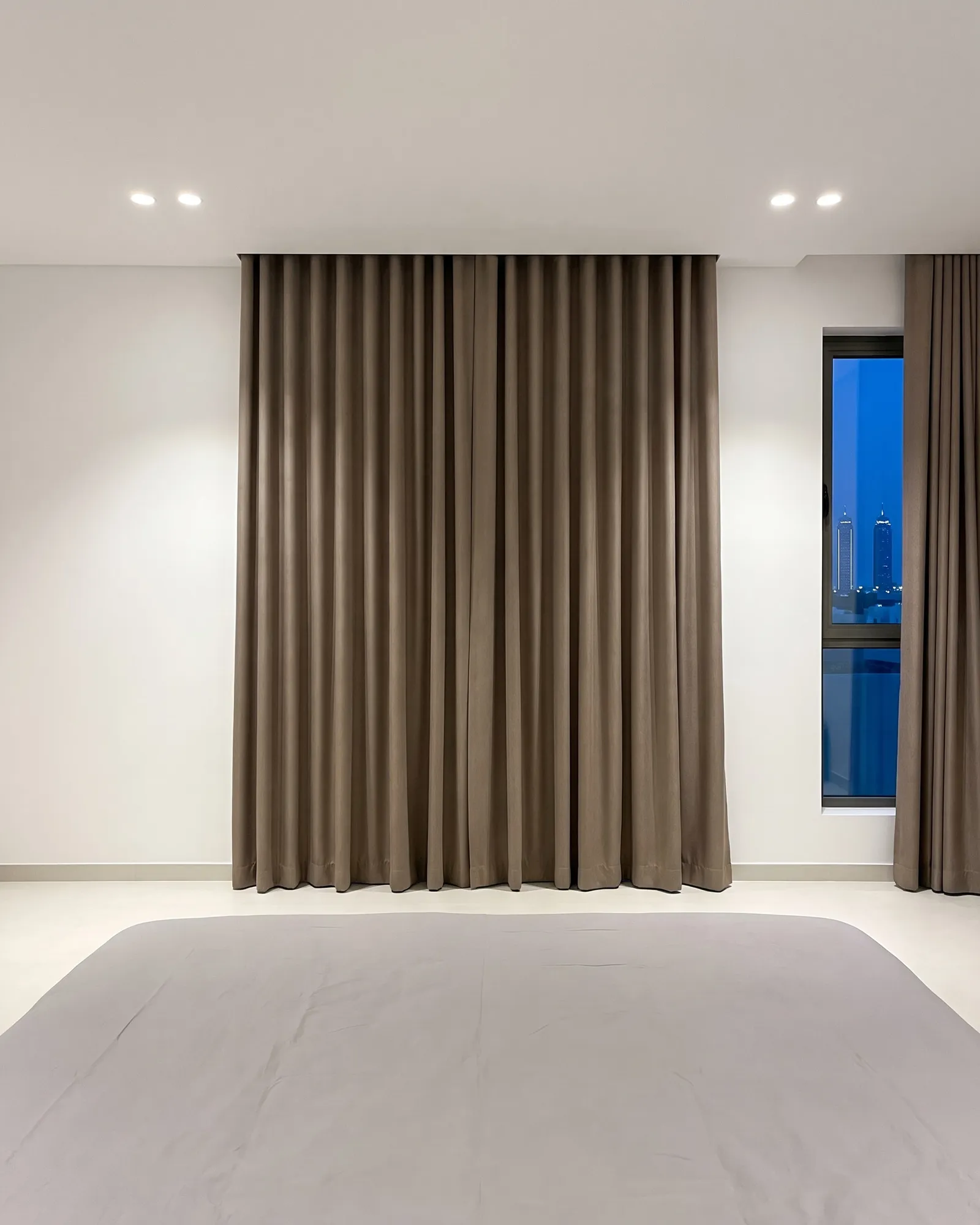 Taupe heavy curtains closed across a bedroom window at dusk, a narrow window beside them with its own curtain drawn open, lit towers