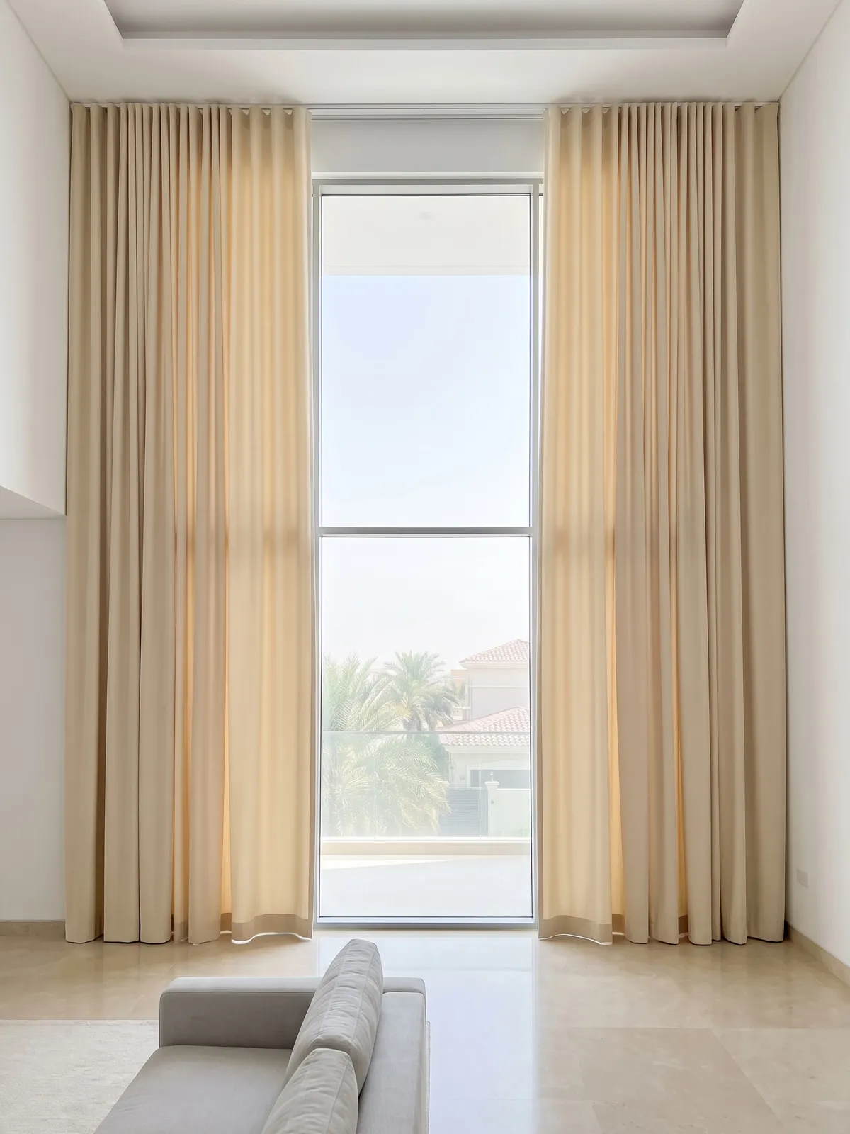 Cream heavy curtains drawn open at both sides of tall living-room glass, a stone floor, a high ceiling and a grey sofa in front