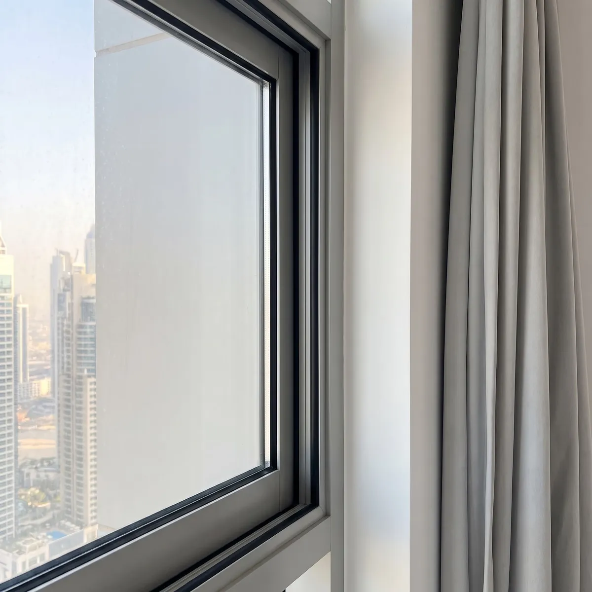 Close view of a shut sliding window's aluminium frame and its seal, towers outside, a light grey curtain hanging at the right edge