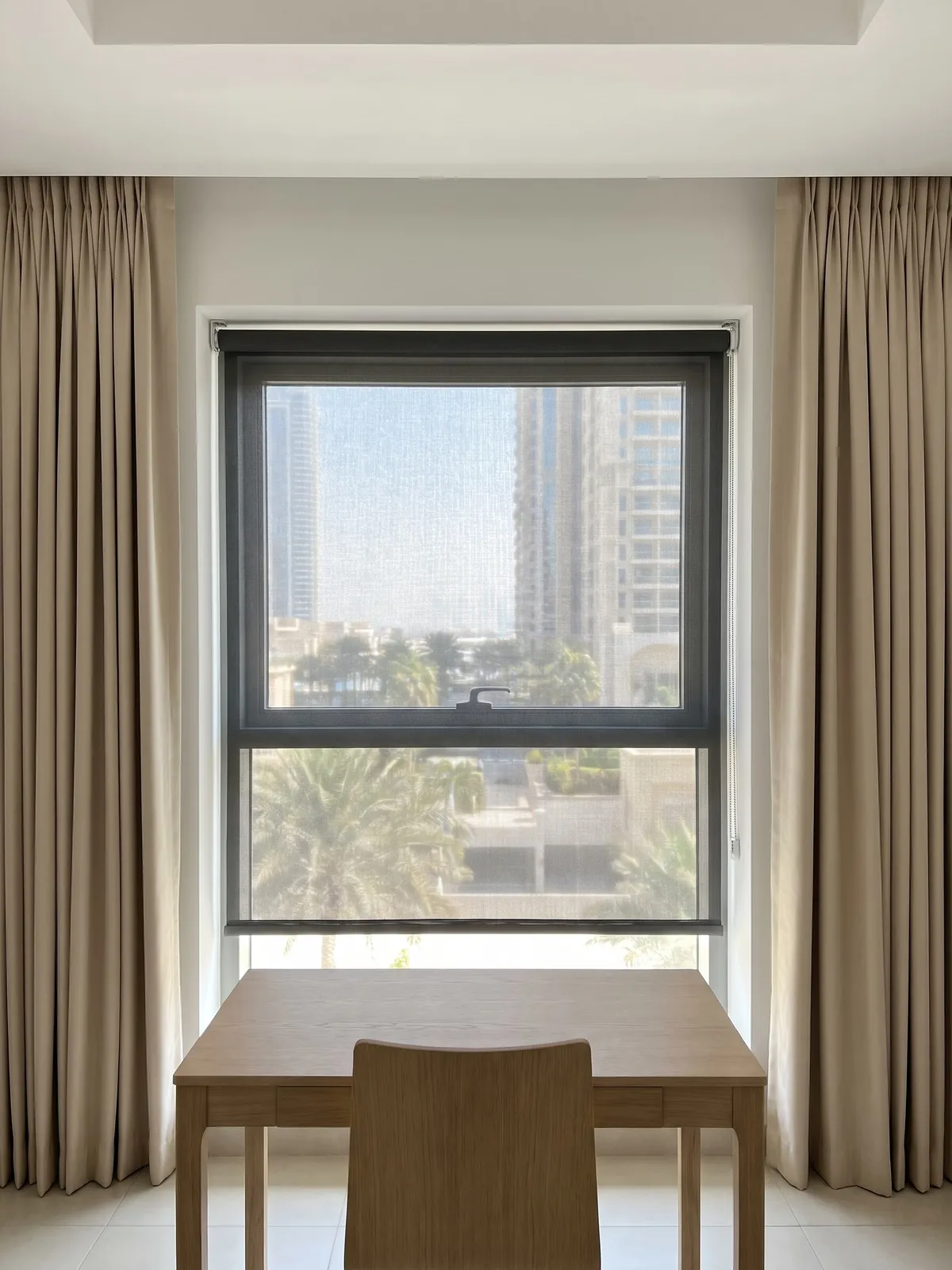 Charcoal sunscreen roller lowered on a window behind a wooden desk, light beige curtains open at both sides, palms and towers through it