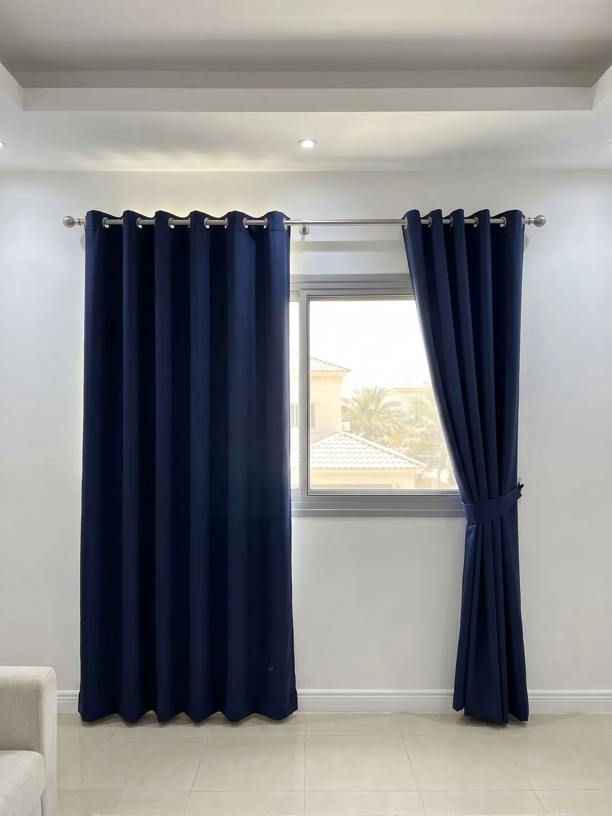 Navy eyelet curtains on a silver wall pole drawn open at both sides of a window, one tied back, palms and villas outside