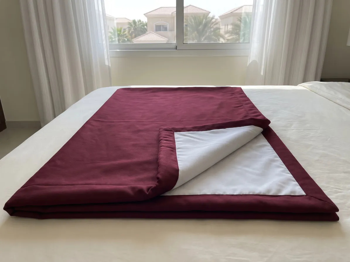 Burgundy curtain folded on a bed with its corner turned back to show the white lining, a window with sheers and villas behind