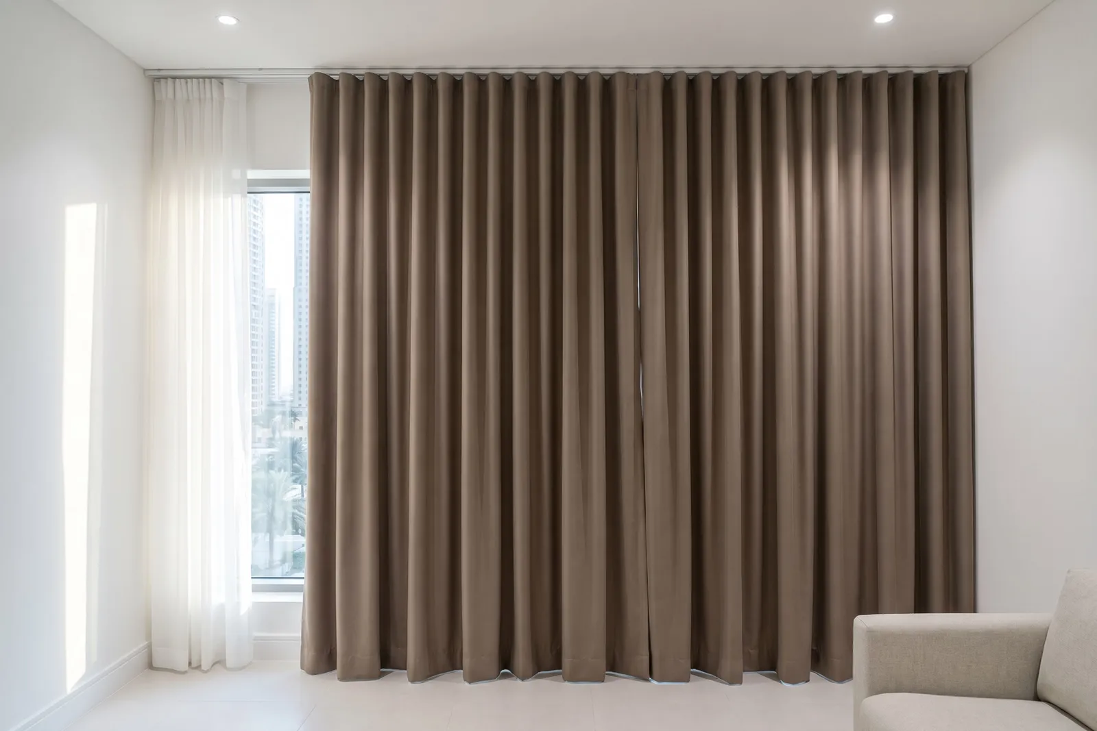 Taupe thermal curtains closed on a ceiling track across west-facing glass, a narrow window beside them with a white sheer, a sofa arm