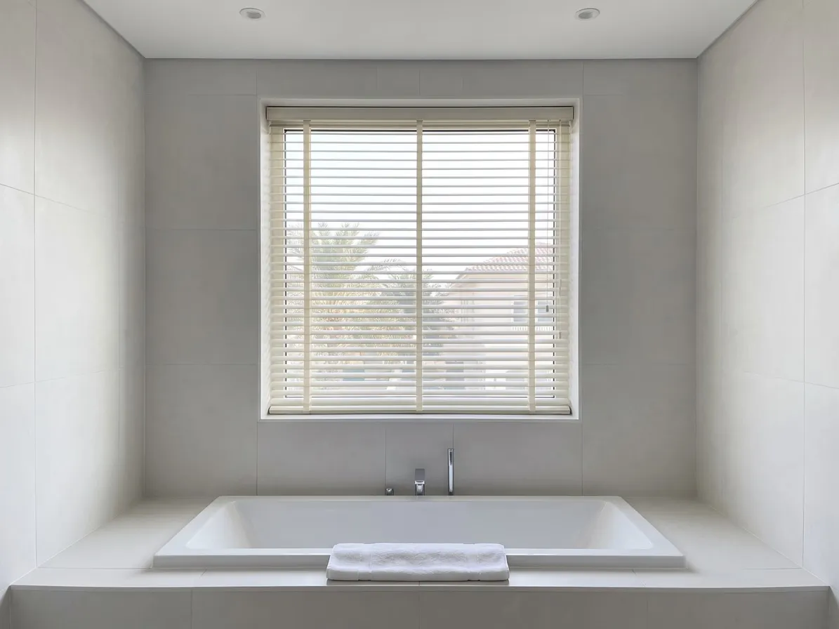 Ivory Venetian blind half open in a tiled bathroom window above a white bath, palms and a villa roof in the gaps, a towel on the bath edge