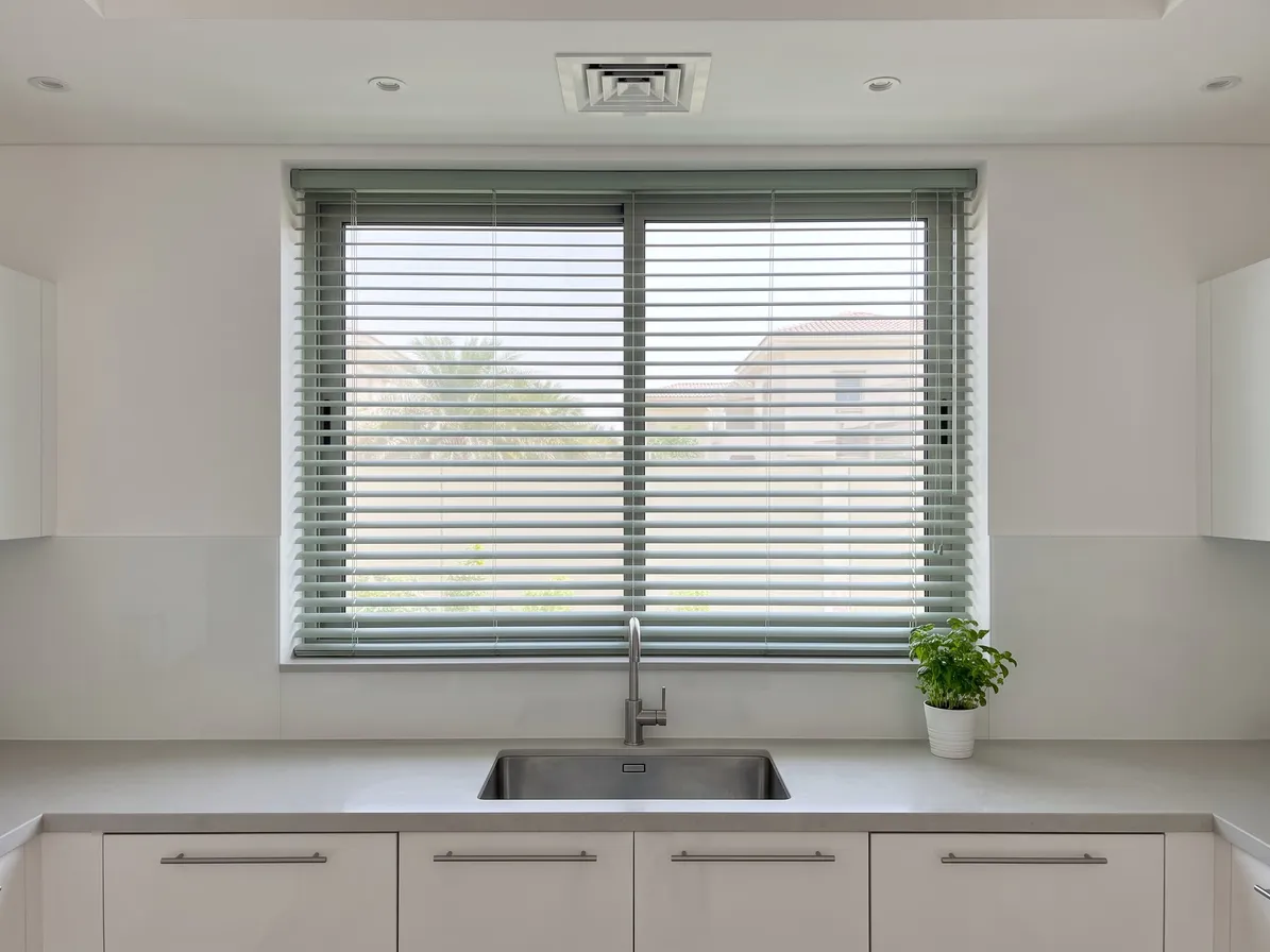 Sage-green Venetian blind open in a two-pane kitchen window above a steel sink and tap, a herb pot on the worktop at the right
