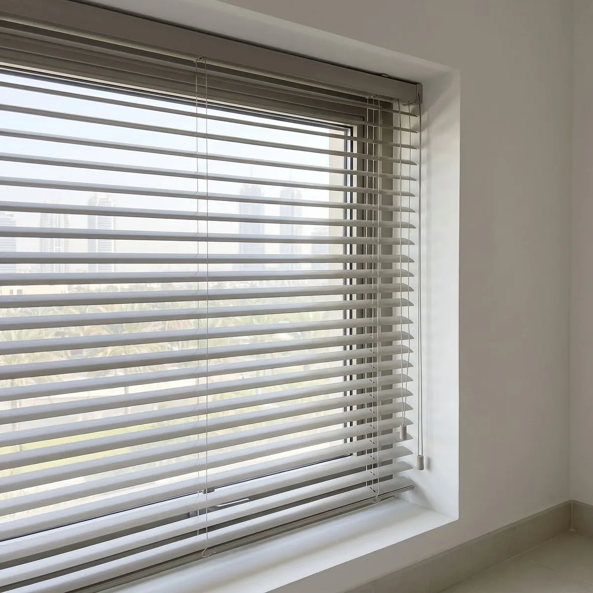 Pale grey Venetian blind seen at an angle with half-tilted slats, two thin cords with small pulls hanging from the right end of the headrail