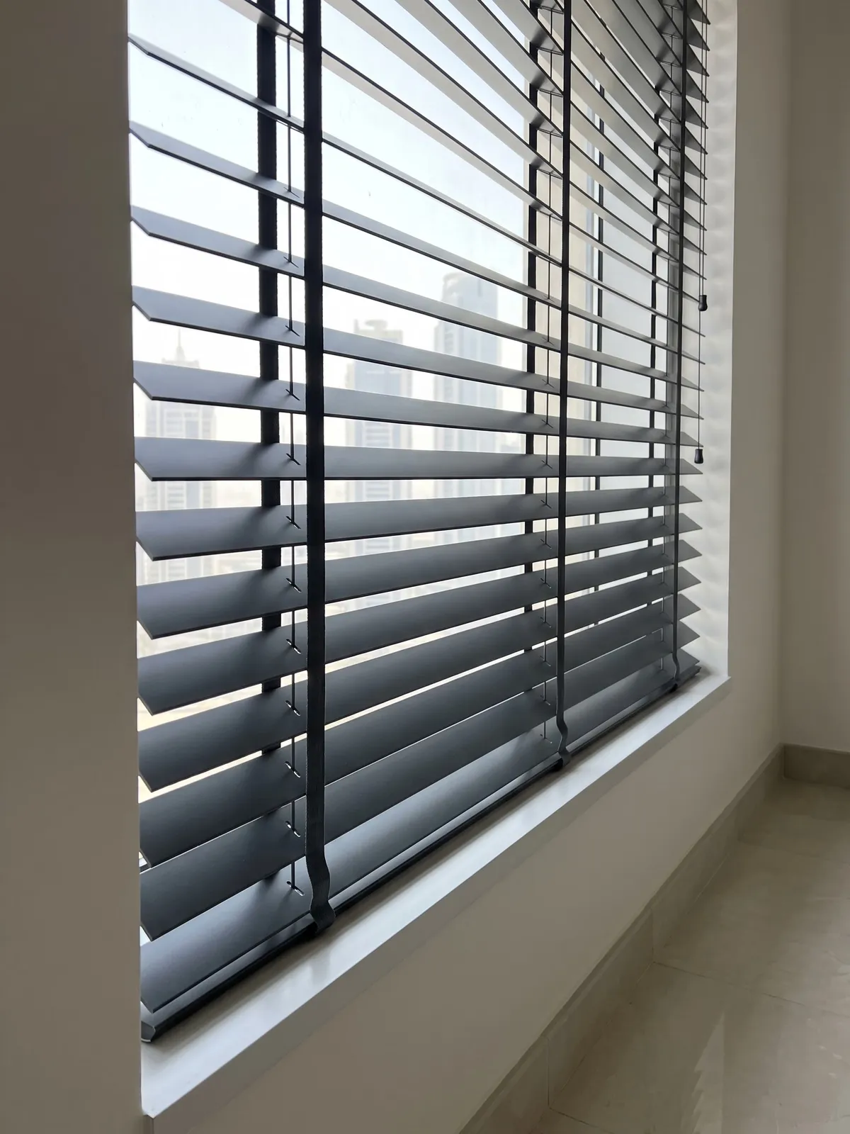 Charcoal Venetian blind seen from the side with half-tilted slats, wide dark tapes down its face and a thin rung string at the near end