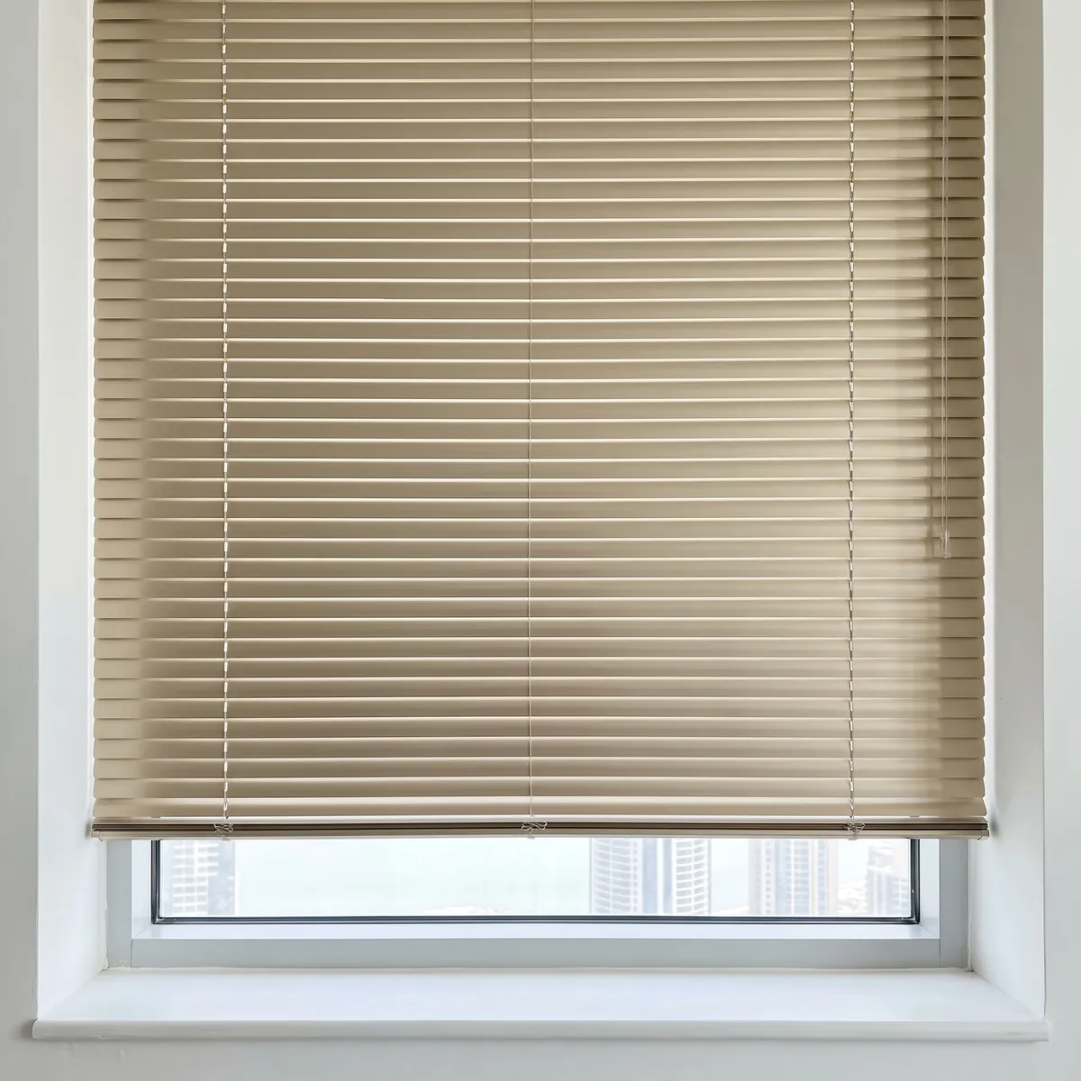 Champagne Venetian blind with closed slats in even level lines above a strip of glass with towers beyond and a white sill below