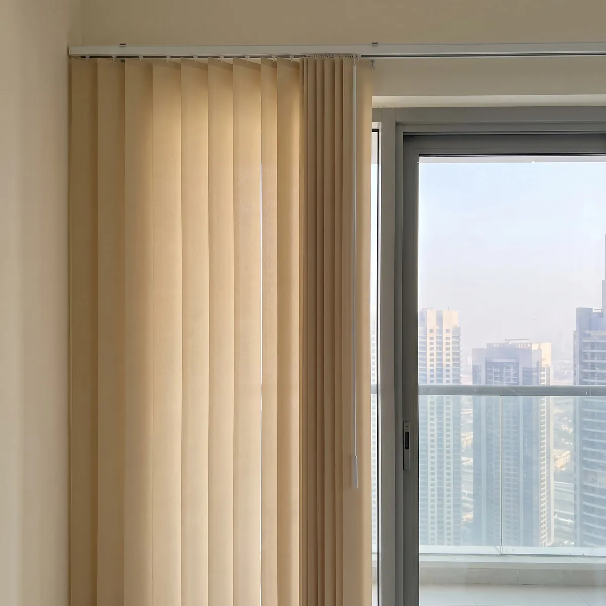 Sand vertical blind with a white control wand hanging in front of its end louvre beside a sliding door with towers beyond