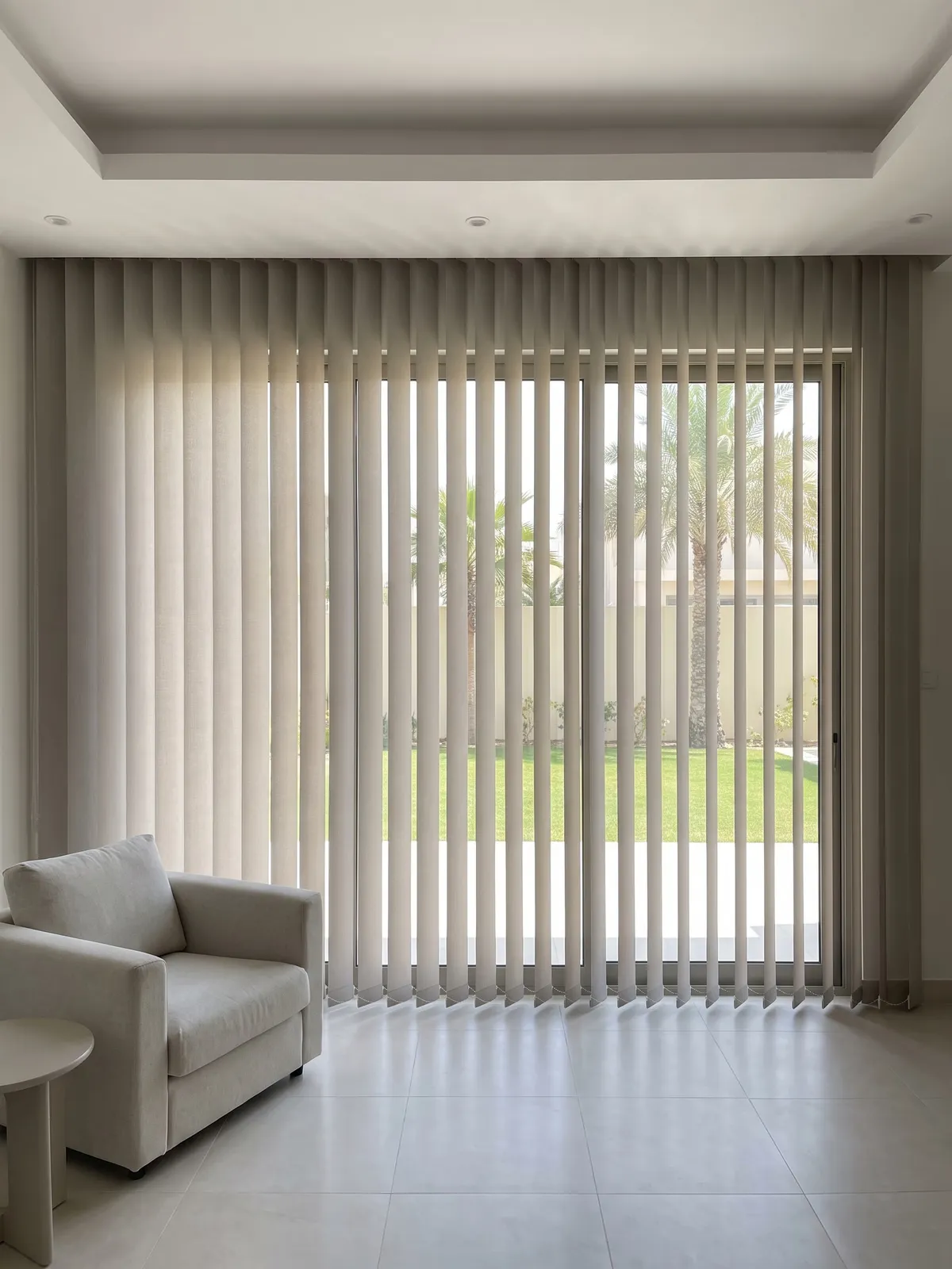 Greige vertical blind across wide garden doors, the right half angled open to show lawn and palms, an armchair at the lower left