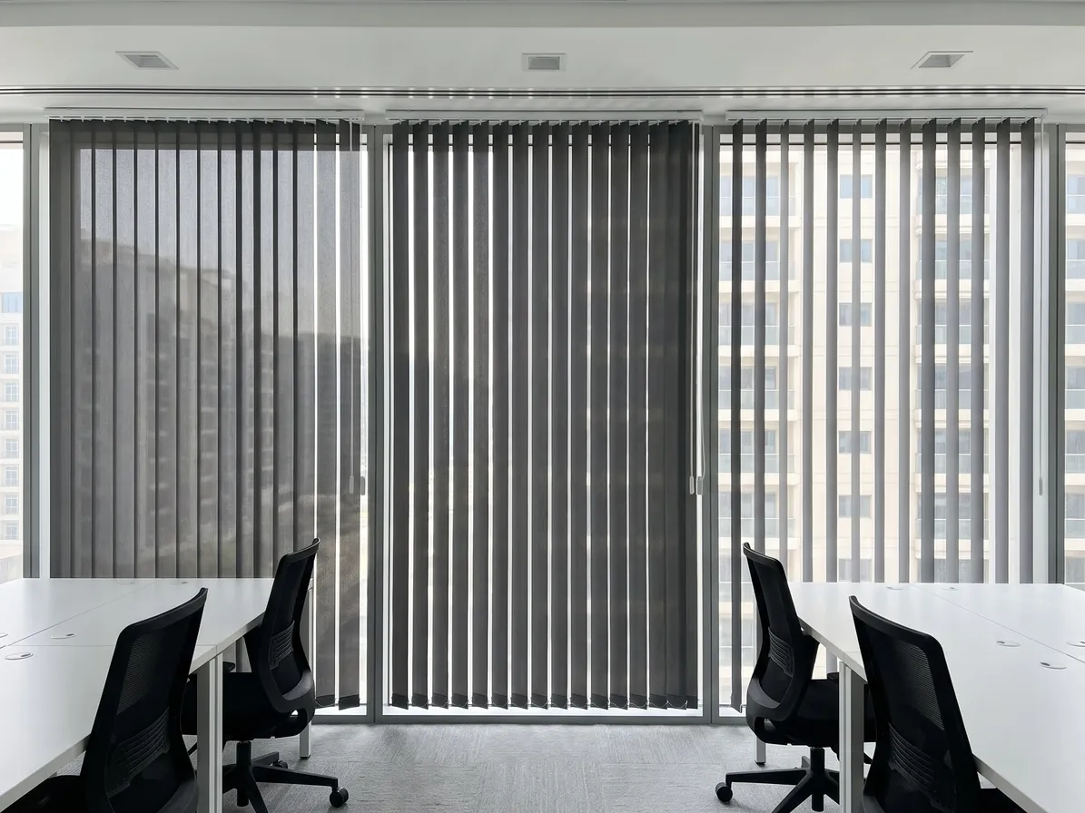 Three charcoal vertical blinds across an office glass wall, the right one turned edge-on to show the buildings, bare white desks and chairs in front