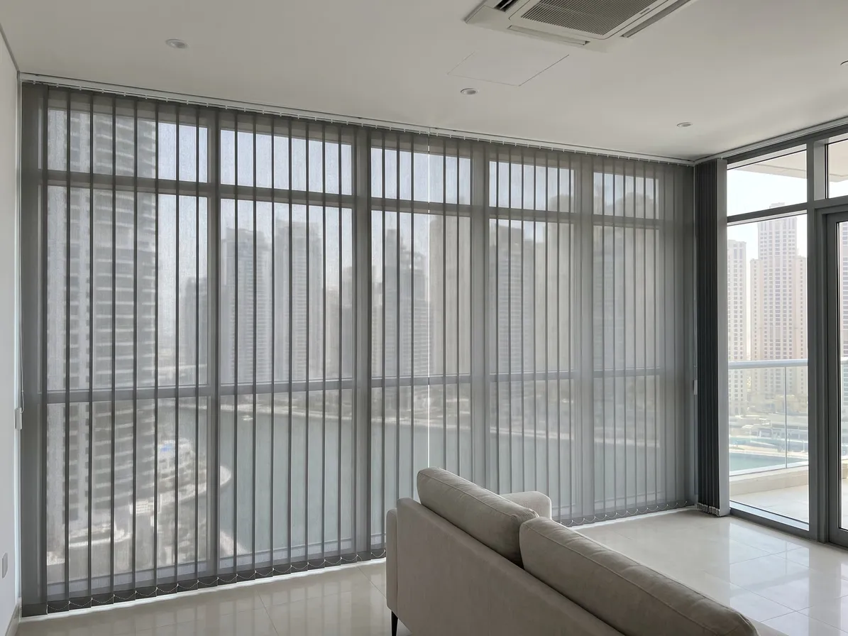 Mid-grey sunscreen vertical blind across a floor-to-ceiling glass wall, towers and water showing through, a glass wall and balcony at the right
