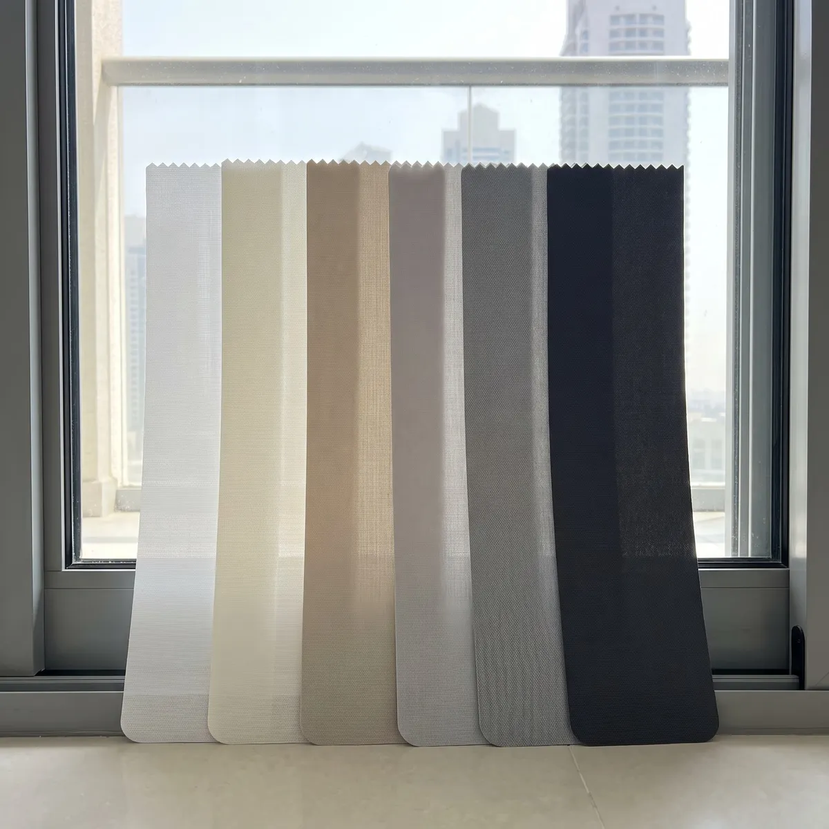 Six vertical-blind fabric samples with zig-zag tops leaning against a balcony door, from white and ivory to charcoal, towers behind