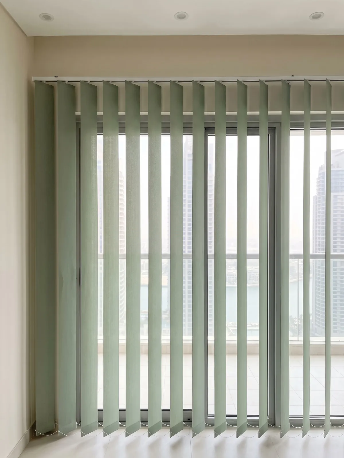 Muted sage vertical blind louvres turned half open across a balcony door, strips of daylight showing the frame, a glass railing and towers