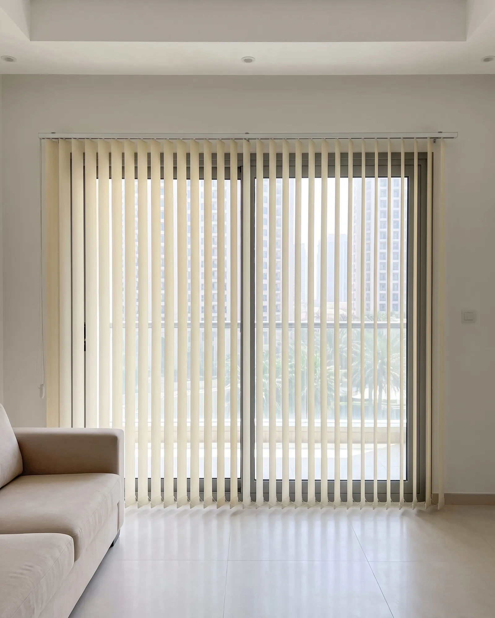 Cream vertical blind across a sliding balcony door, louvres half turned so daylight, a railing and palm tops show between them, a sofa at the left