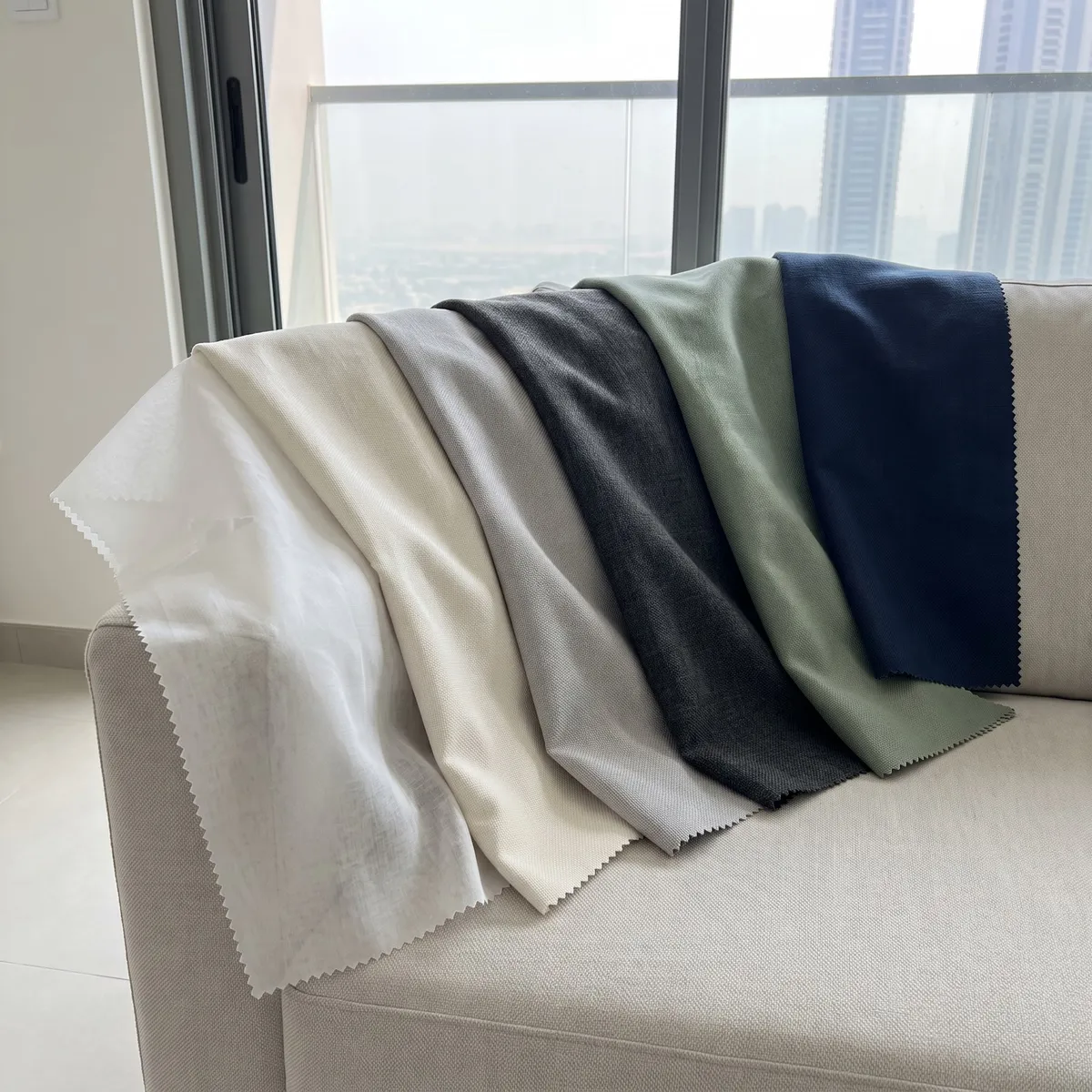 Six fabric samples with zigzag edges draped over a sofa by a balcony door, in white sheer, ivory, grey, charcoal, sage and navy