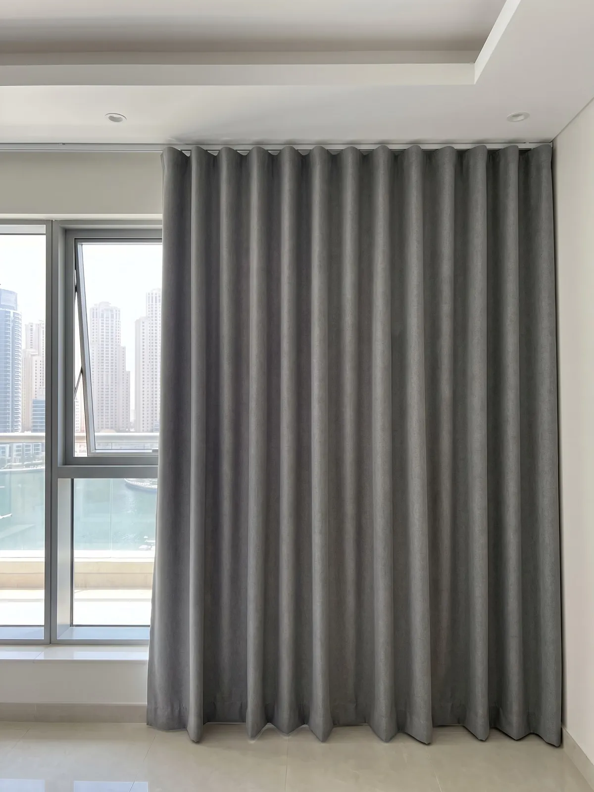 Mid-grey wave curtain closed beside a window with a marina view, deep even curves at the top easing to softer folds at the hem