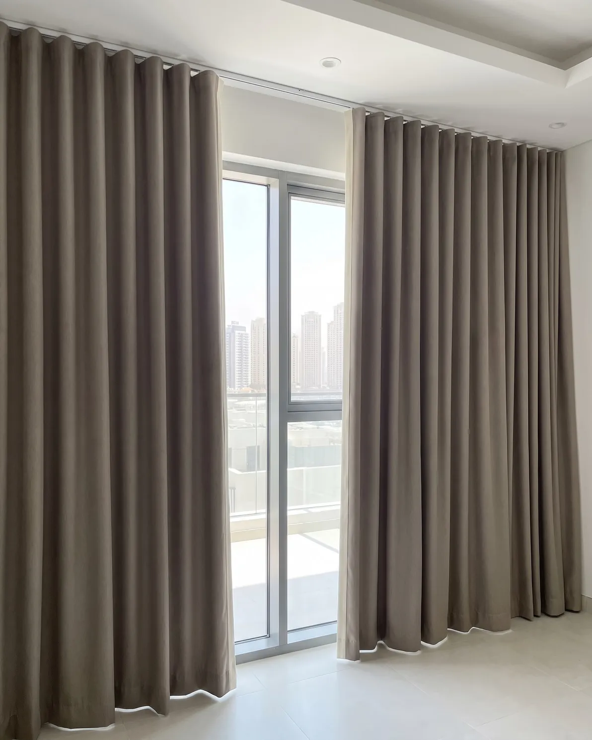 Taupe wave curtains drawn open either side of a tall window, a cream lining showing along each inner edge