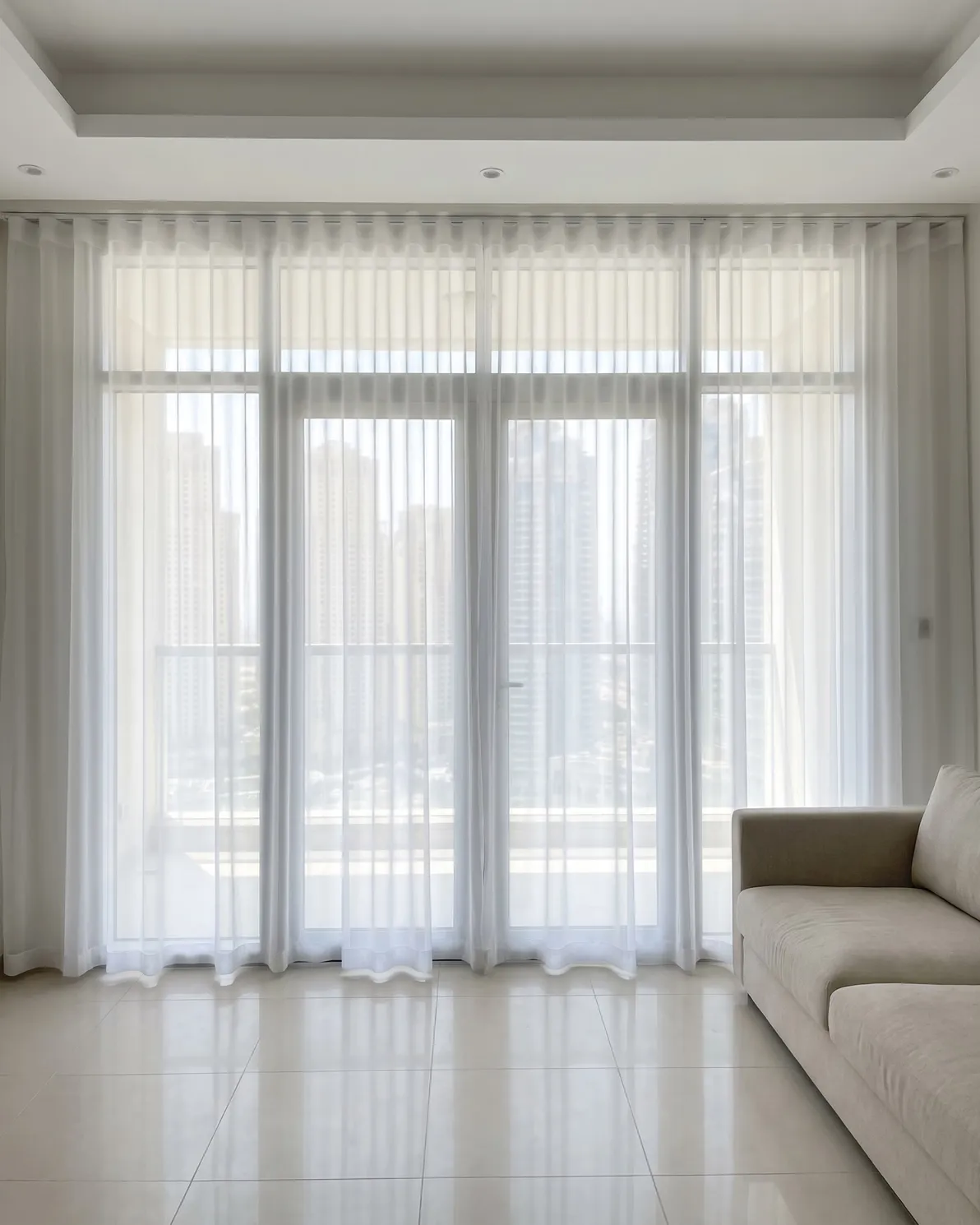 White sheer wave curtains closed across tall glass doors, daylight and towers softly visible through the cloth, a sofa at lower right