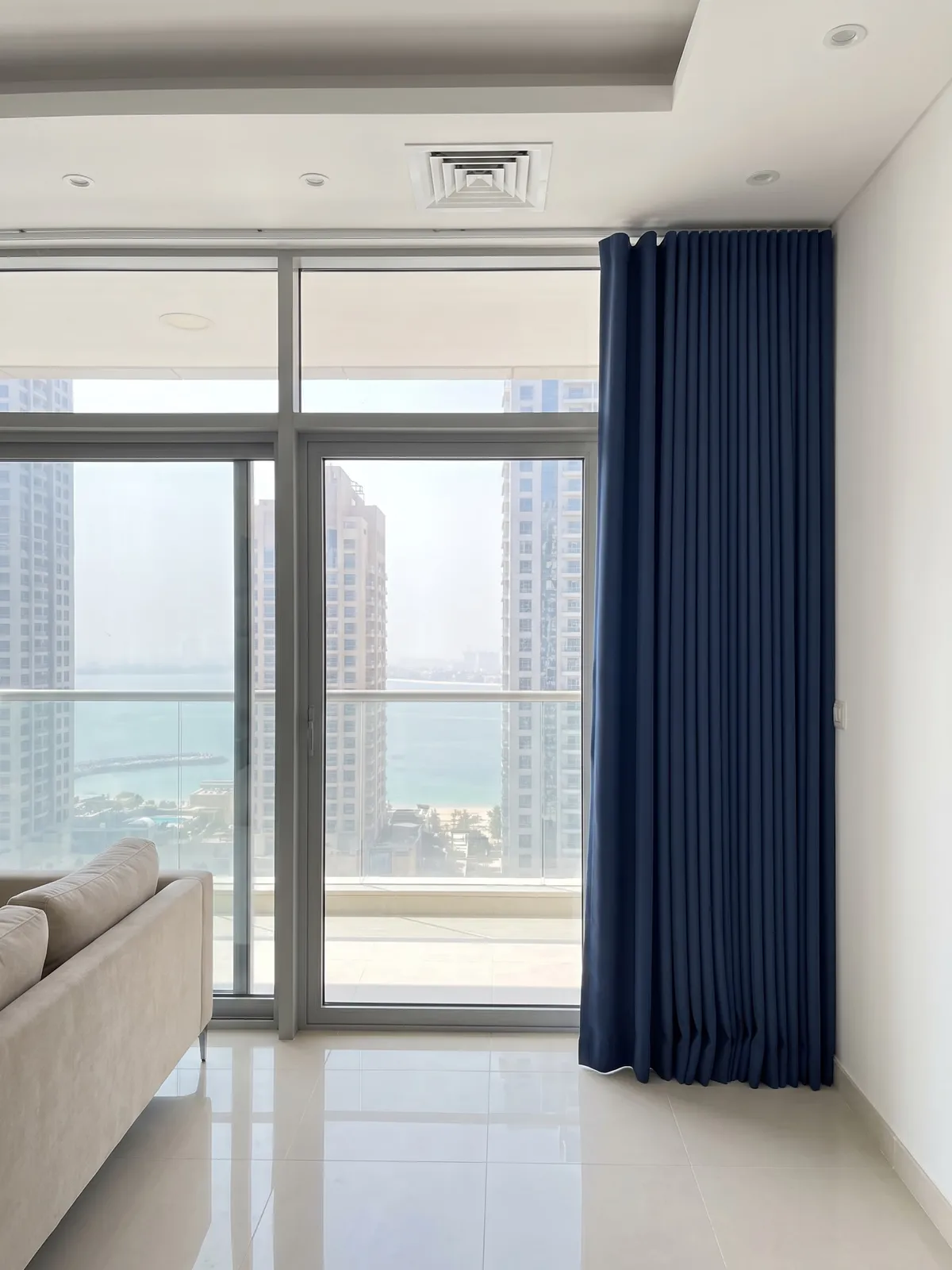 Navy wave curtain drawn fully open and stacked against the right wall beside tall glass doors with a sea view, a sofa at lower left